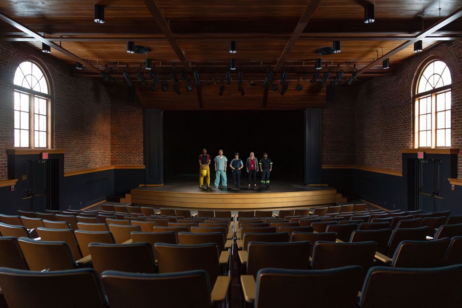 Group of performers standing on stage in an empty theater auditorium with wooden seats and arched windows.