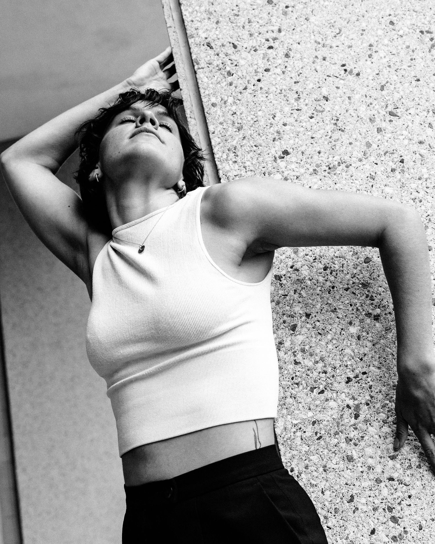 A person wearing a white crop top leans back against a textured wall in dramatic black and white composition.