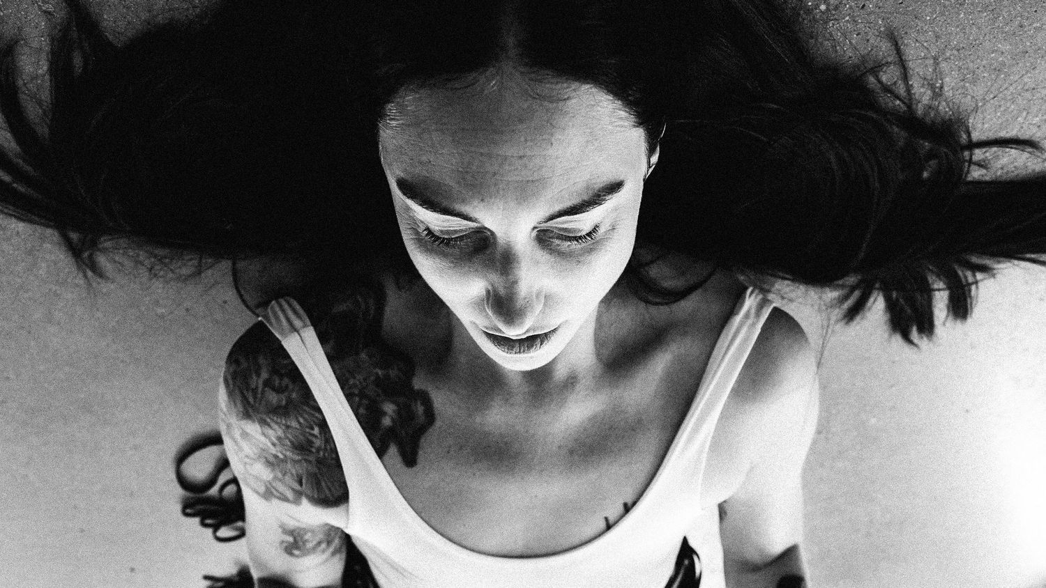 A dramatic black and white portrait showing a figure with dark hair and intense lighting leaning forward in a white top.