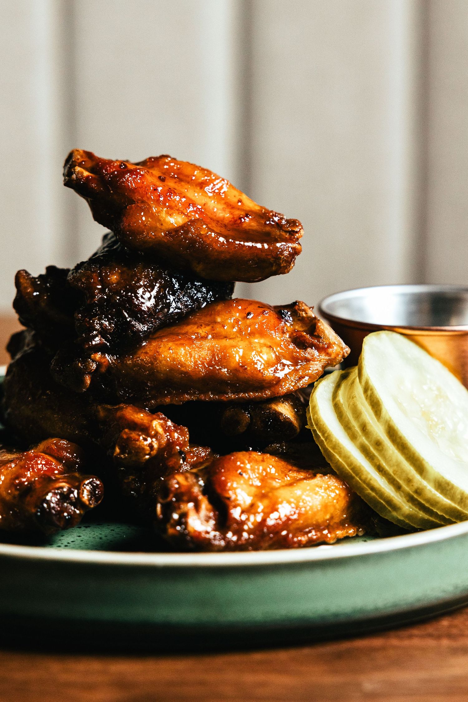 Glazed chicken wings stacked on a green plate with sliced pickles on the side in moody lighting.
