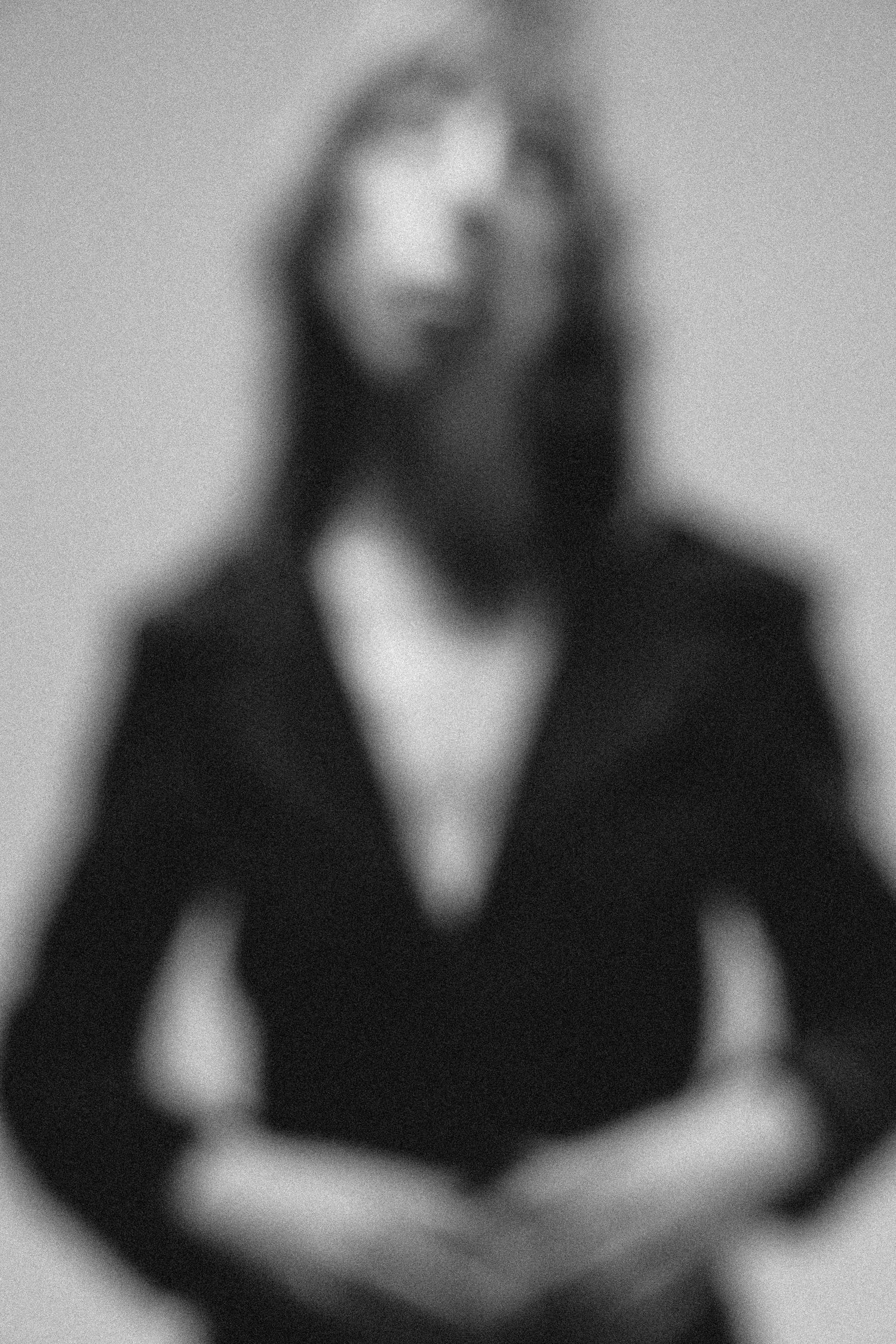 Artistic black and white portrait with intentional blur effect showing figure in dark blazer against plain background.