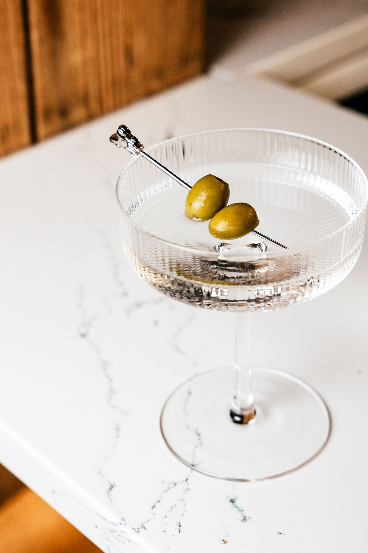 A crystal martini glass with two green olives on a cocktail pick sits on a white marble surface.