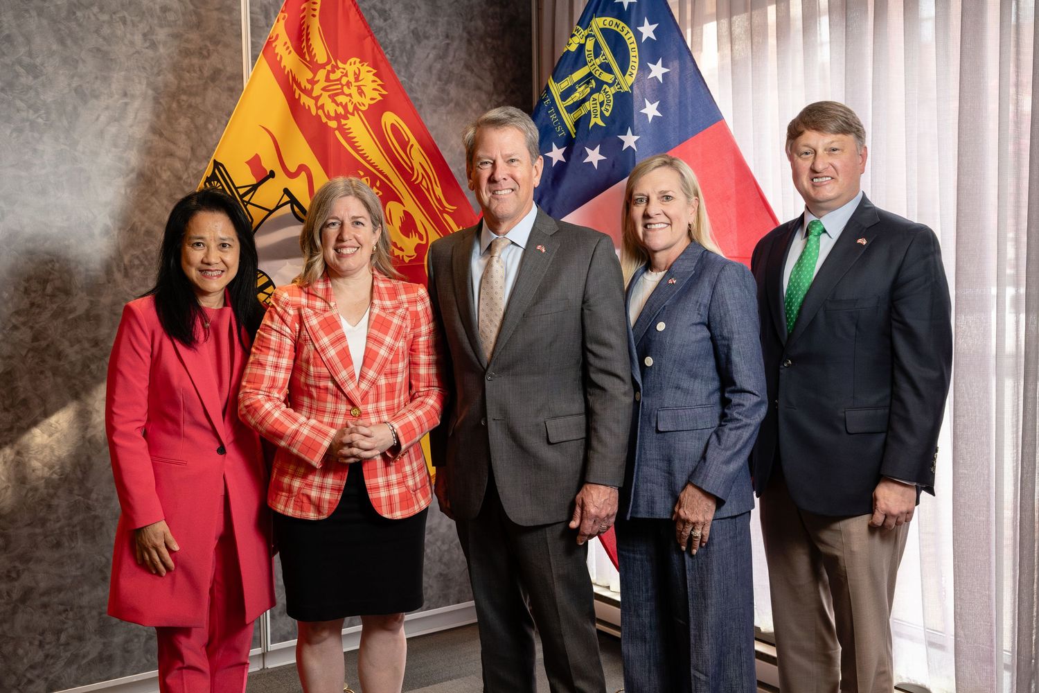 Politicians and officials stand together at a professional event wearing business attire against a gray wall.