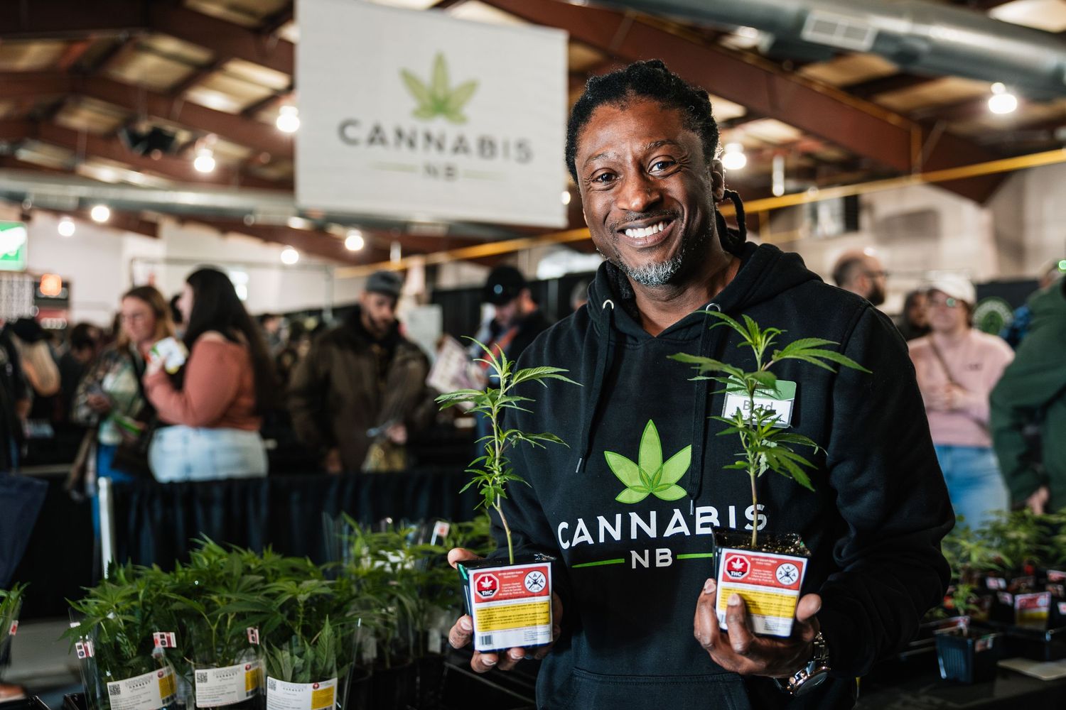 A vendor displays cannabis plants and products at a busy indoor marketplace.