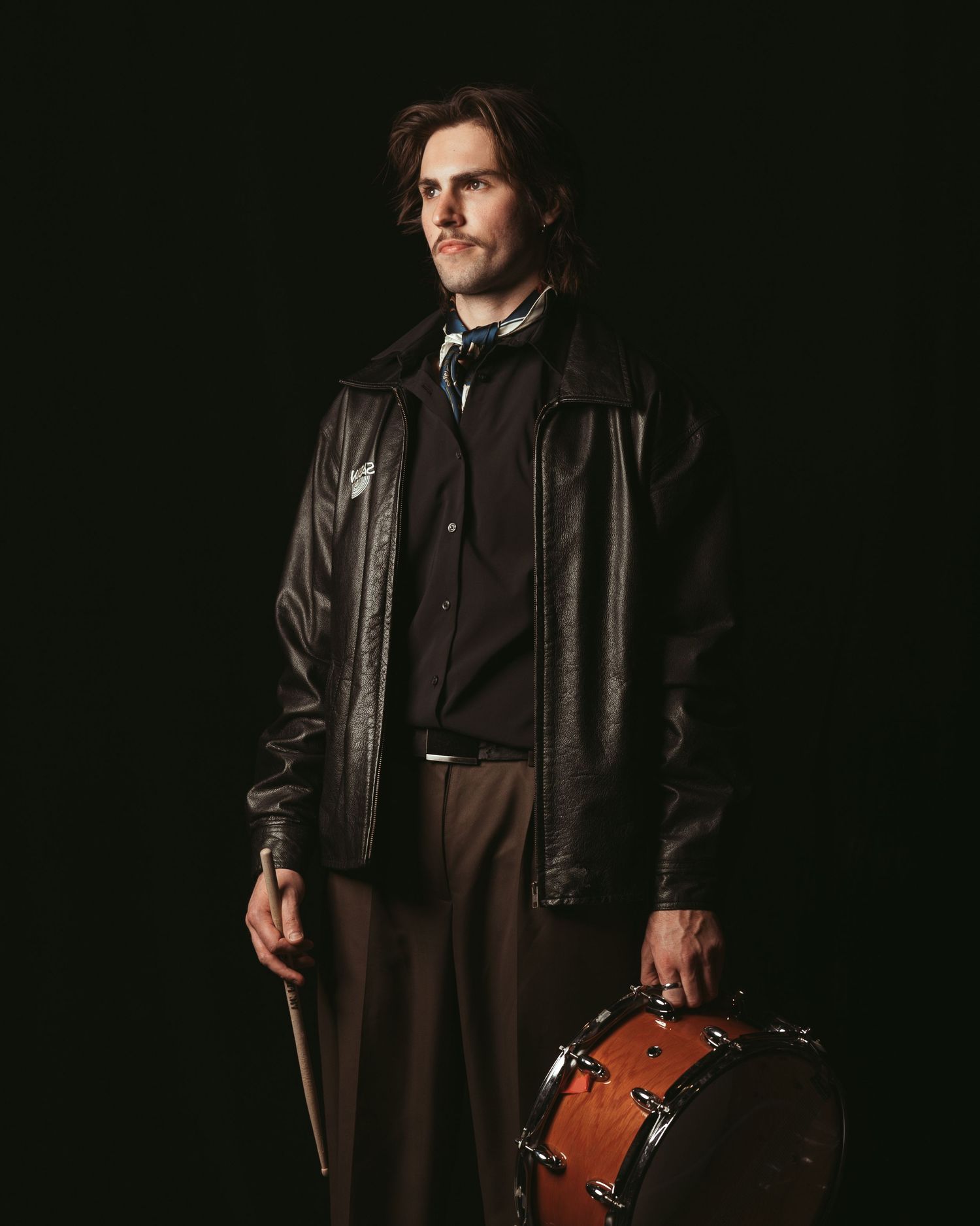 Portrait of a musician in vintage-style attire with a brown leather jacket standing next to a drum in a dark studio.