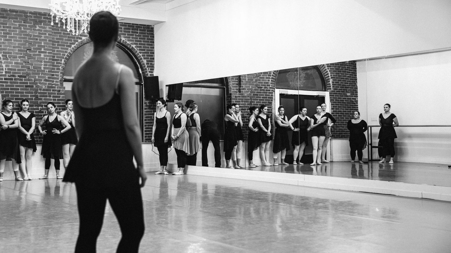 A dance instructor leads a group class in a mirrored studio with brick walls as students follow along in black attire.