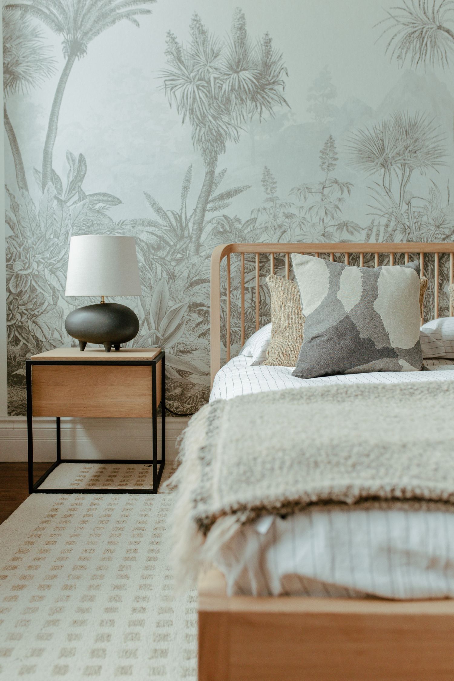 Modern bedroom interior with wooden bed frame, tropical wallpaper mural, and black table lamp on minimalist nightstand.