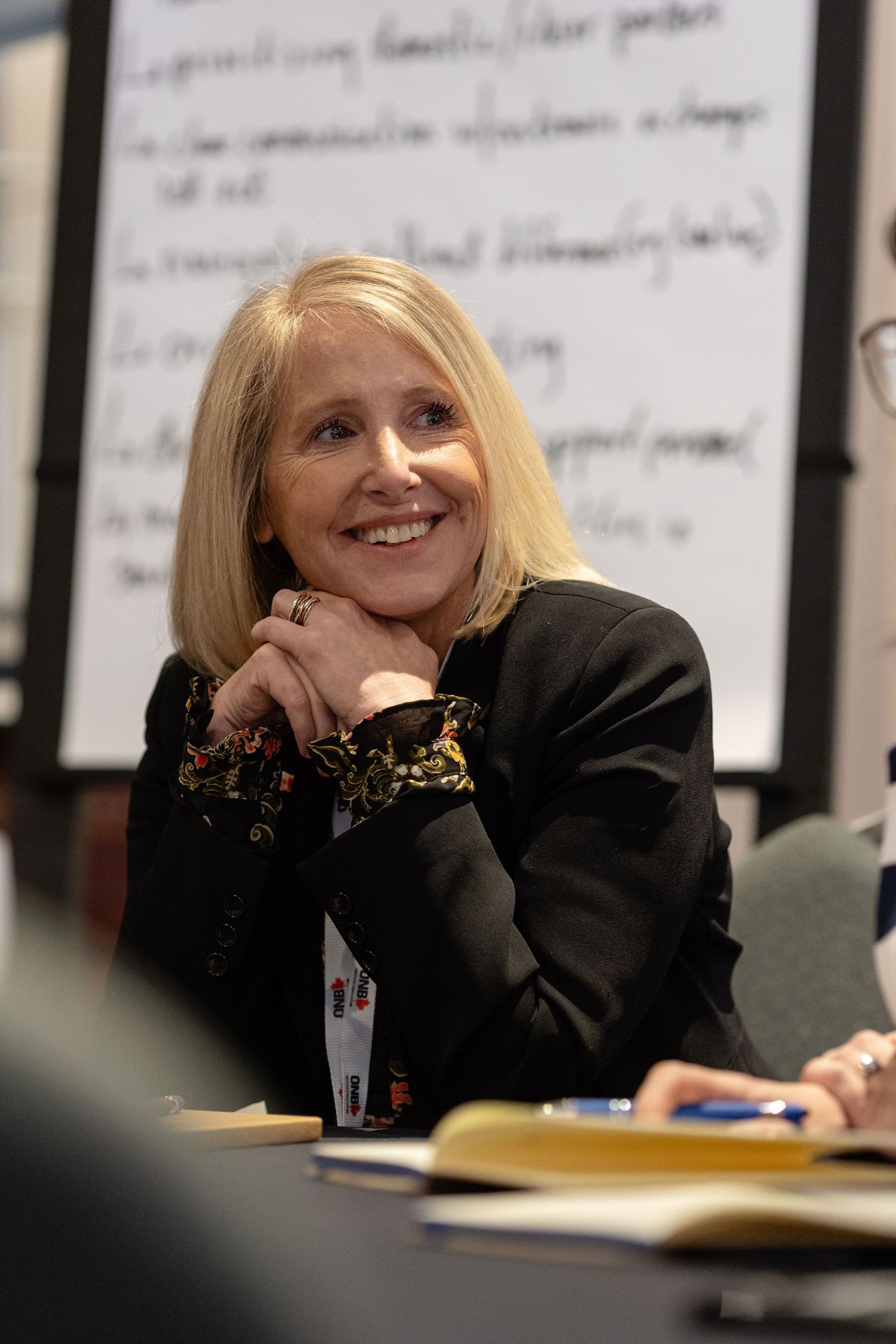 A person with blonde hair smiles warmly while sitting at a desk in an office setting with handwritten notes on the wall.