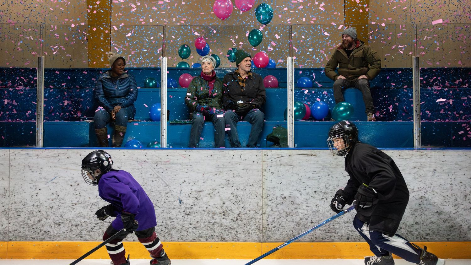 Two hockey players in black and purple jerseys practice drills on an ice rink beneath colorful floating balloons.
