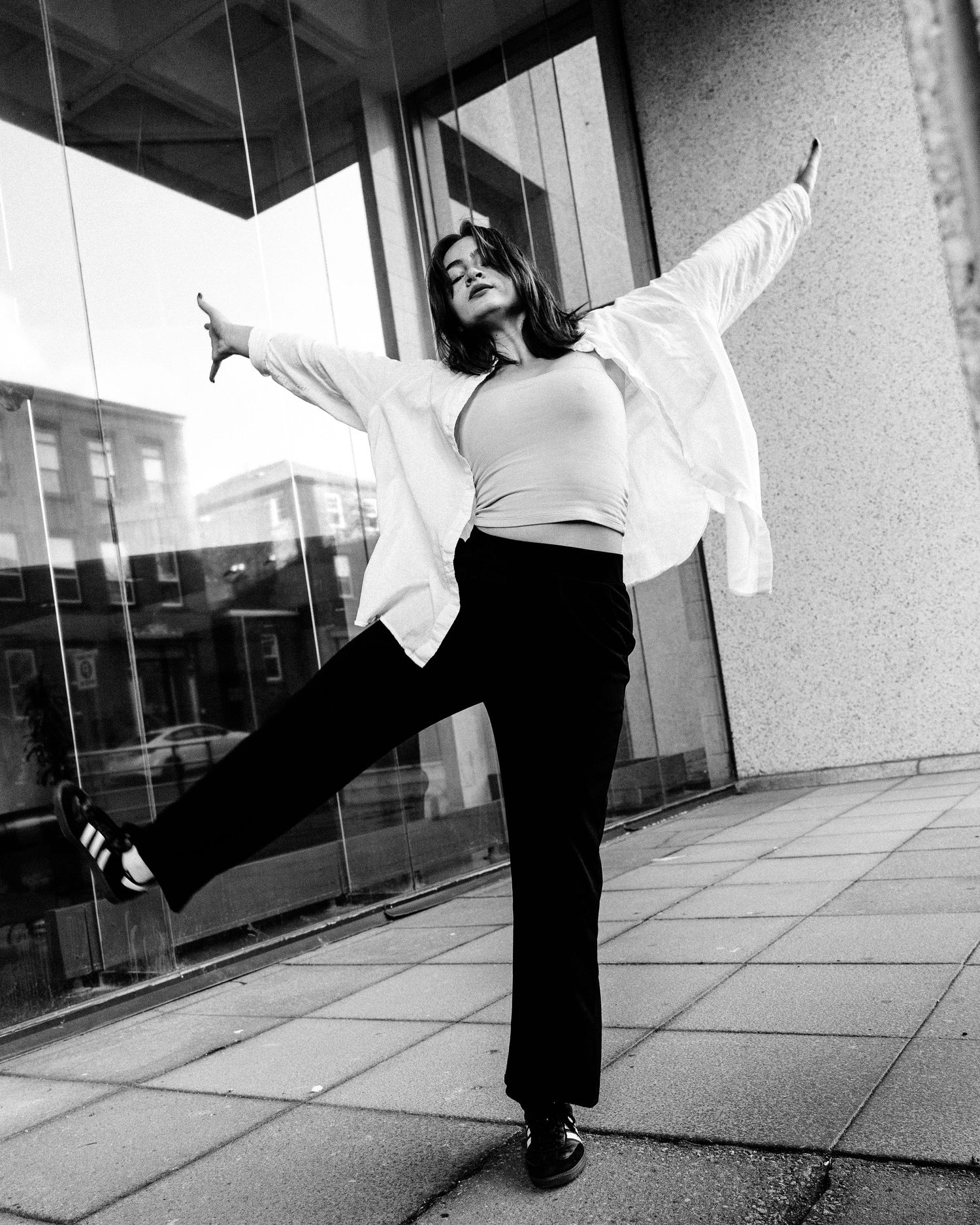 Black and white photo of a person dancing joyfully outdoors wearing a white shirt and black pants in front of a building.