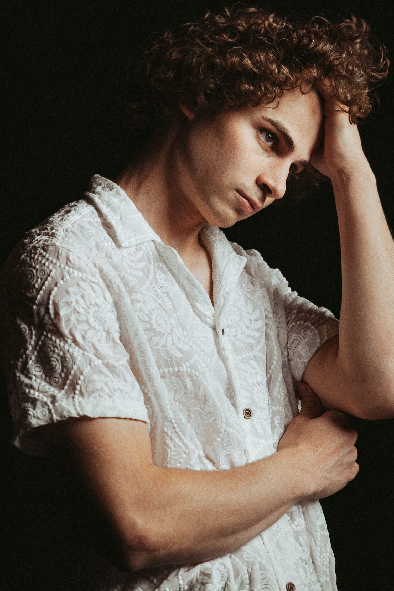 Portrait against dark background of person in white textured button-up shirt with curly hair in contemplative pose.