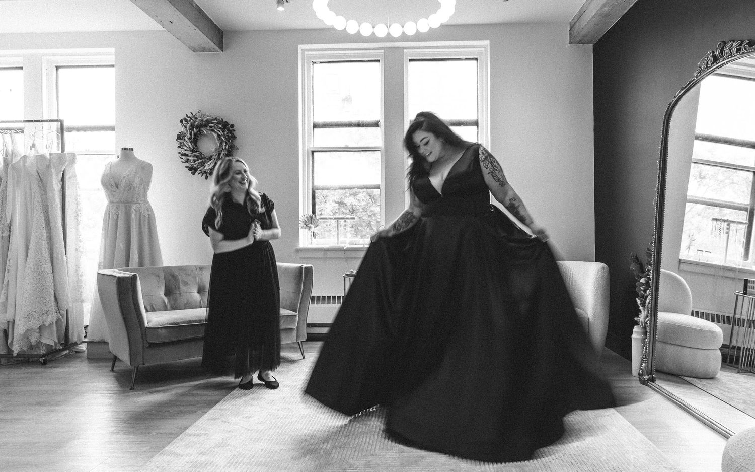 Two people in elegant black dresses move gracefully in a sunlit room with white dresses hanging in the background.