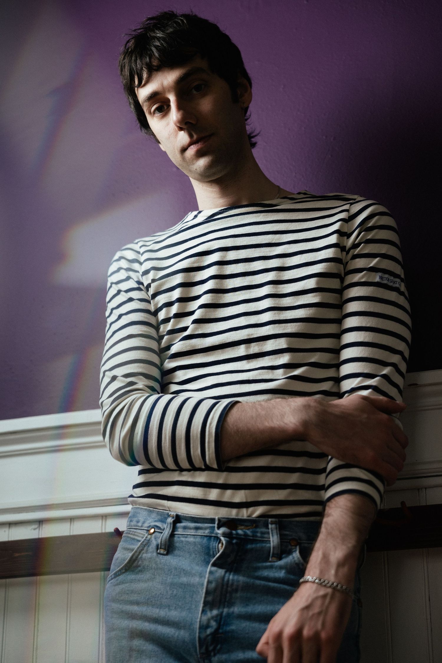 Artistic portrait against purple wall showing person in striped long-sleeve shirt and jeans leaning on white wainscoting.