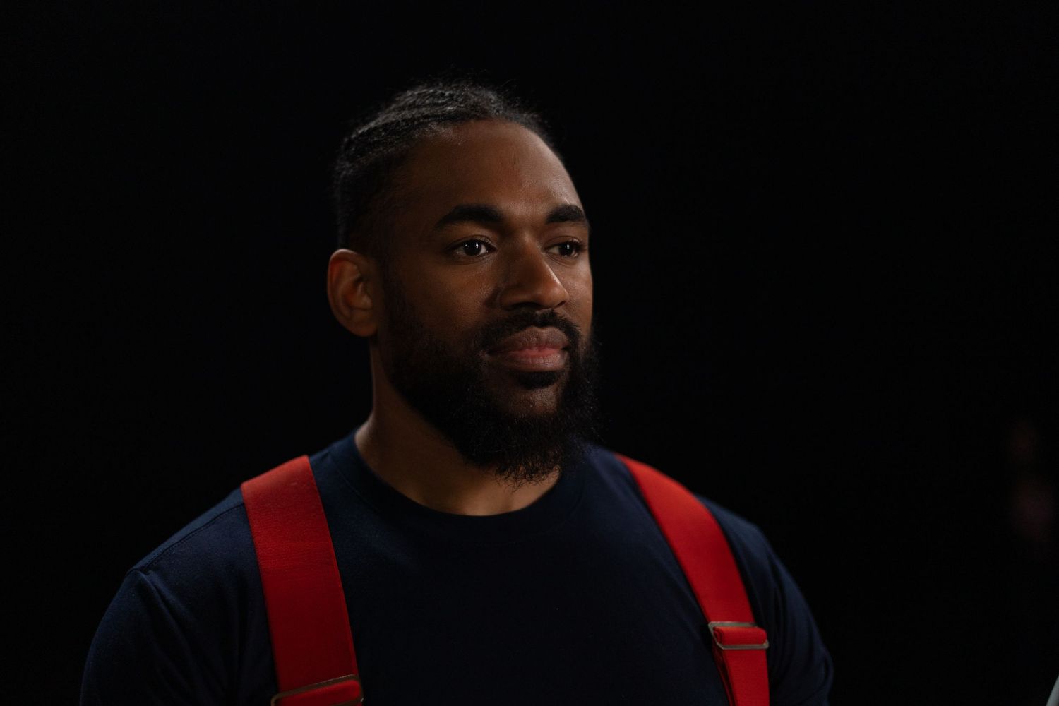 Portrait against dark background of man wearing black t-shirt and red suspenders.