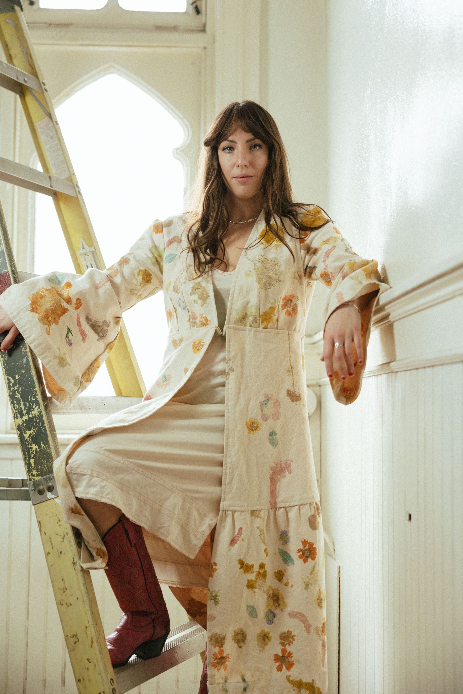 A person poses on a ladder wearing a floral kimono-style robe and burgundy boots in a sunlit room.