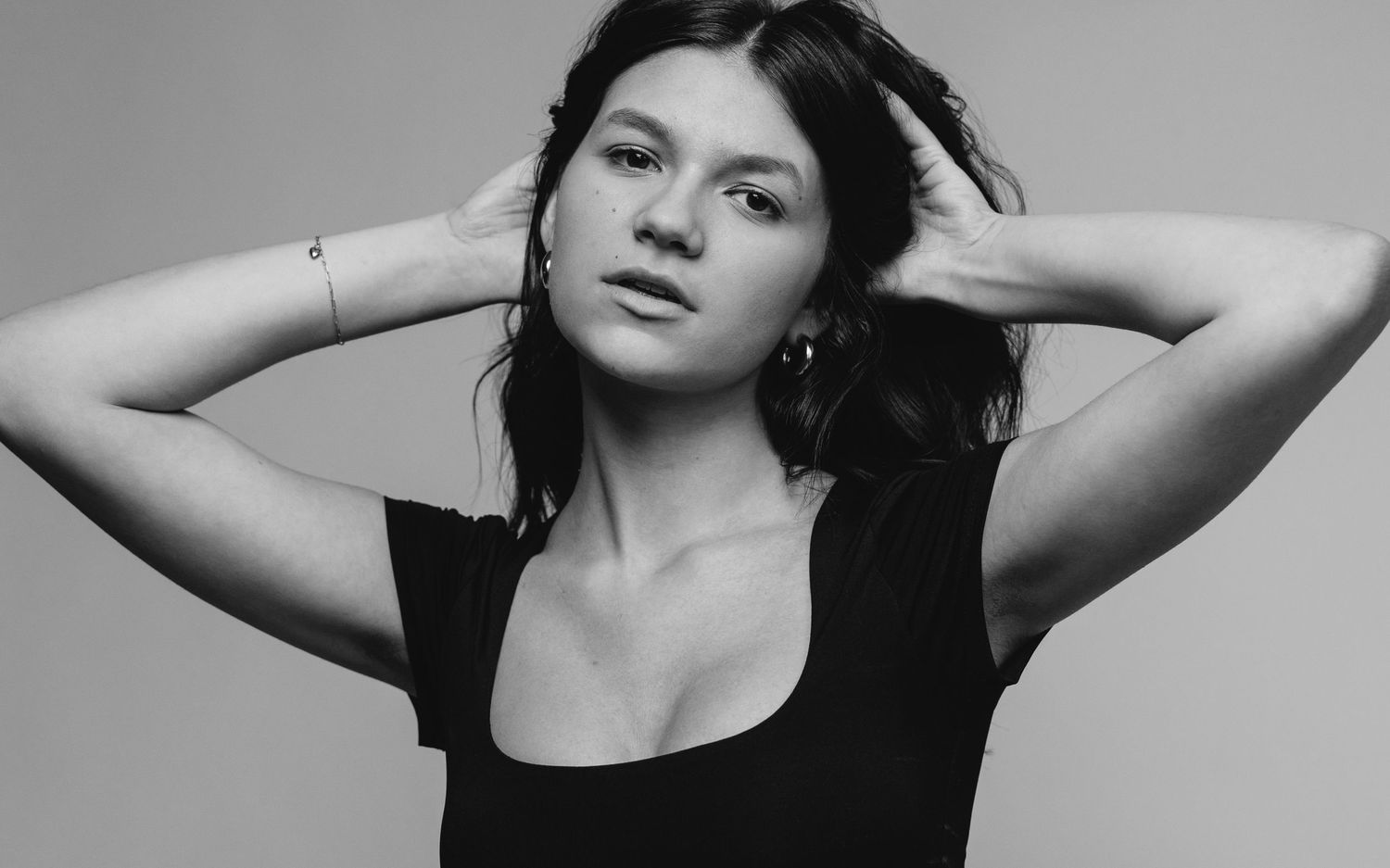 Black and white artistic portrait of a woman in a dark sleeveless top posing with hands behind her head.