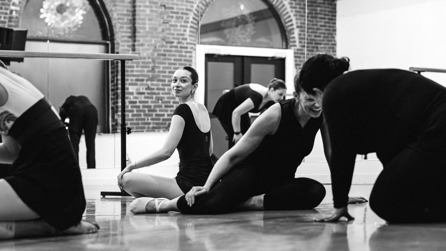 Dancers in black leotards practicing floor movements together in a mirrored studio with brick walls.
