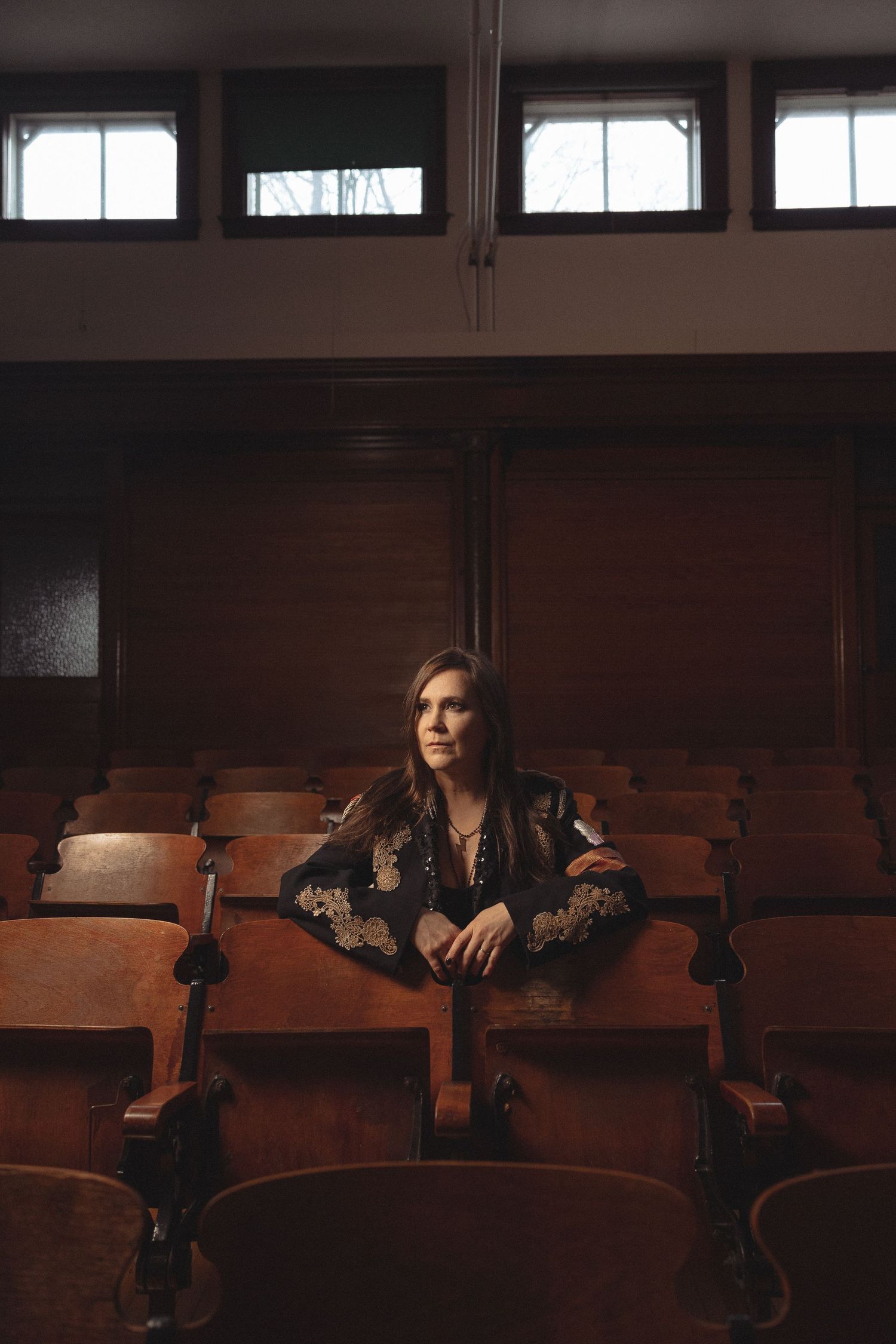 A figure sits alone in vintage wooden theater seats wearing a dark embroidered outfit in moody lighting.