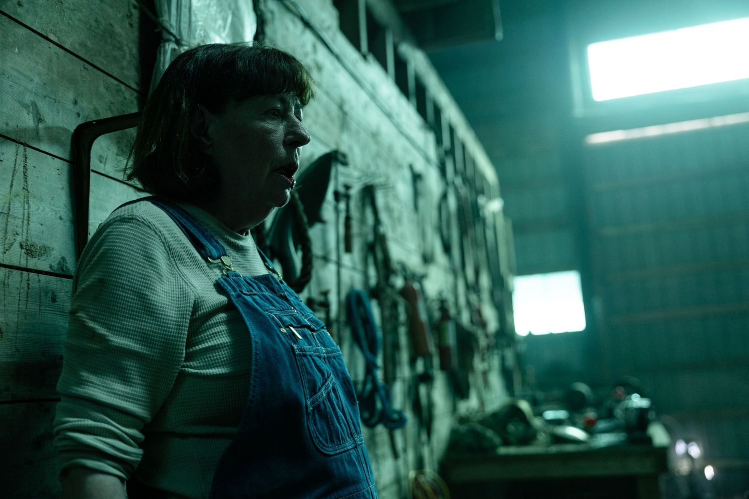 Figure in overalls stands in a dimly lit industrial space with turquoise lighting casting an eerie glow.