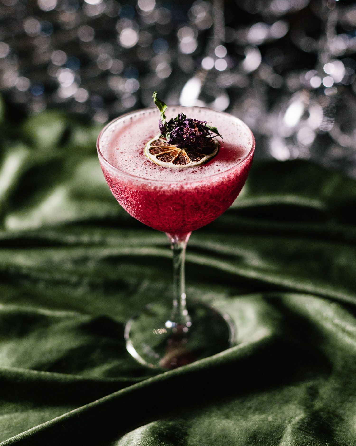 A red cocktail in a coupe glass garnished with dark berries, resting on a green velvet surface with sparkly bokeh background.