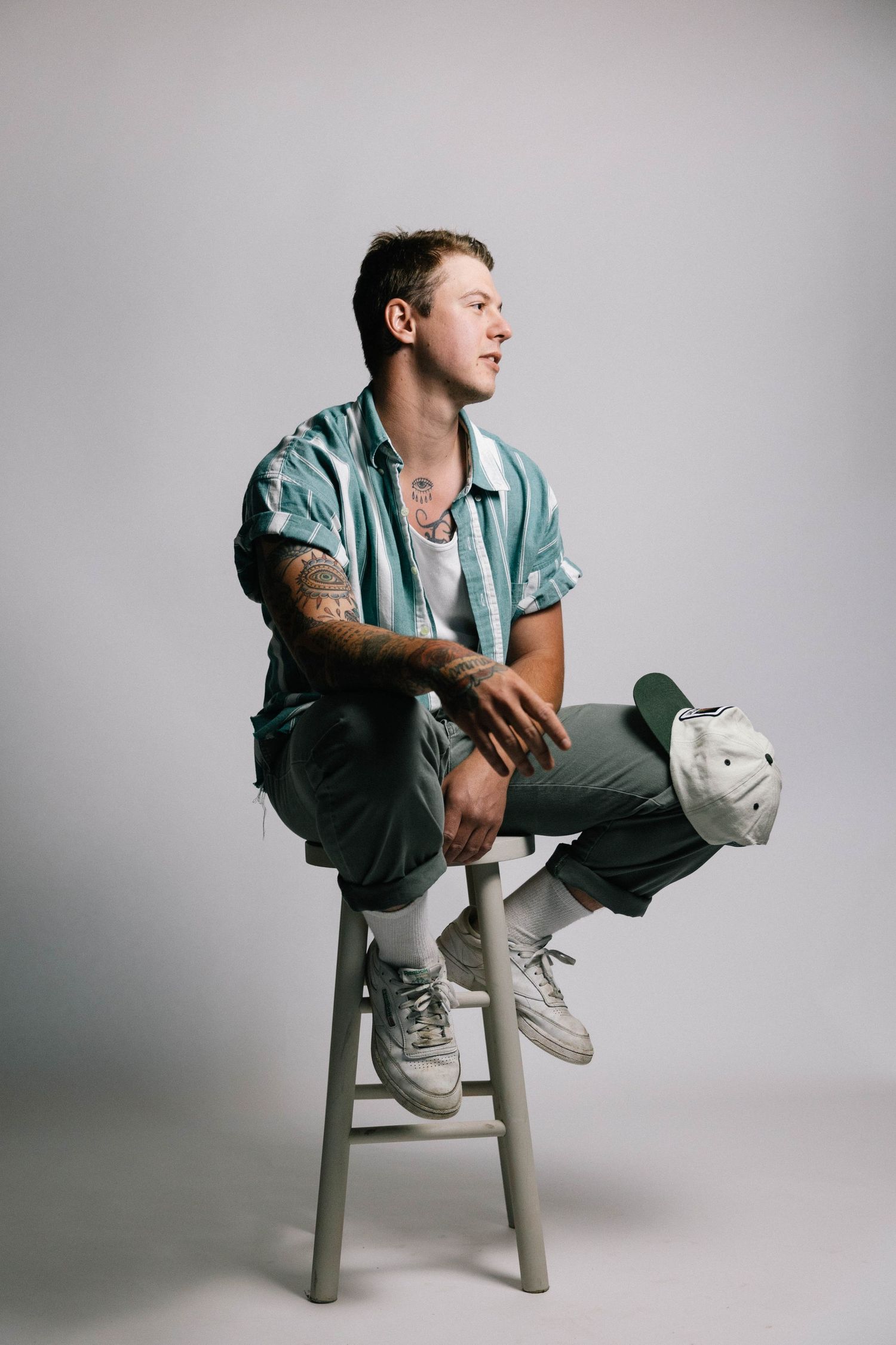 Person in casual mint shirt and pants sits sideways on wooden stool against white backdrop in studio setting.