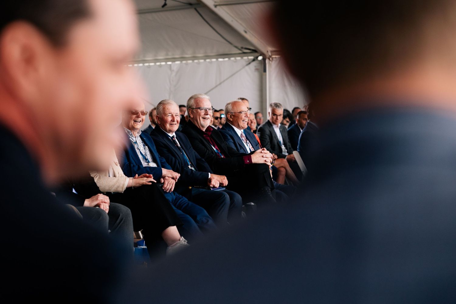 People seated in a row at a formal event viewed through a crowd silhouette in the foreground.