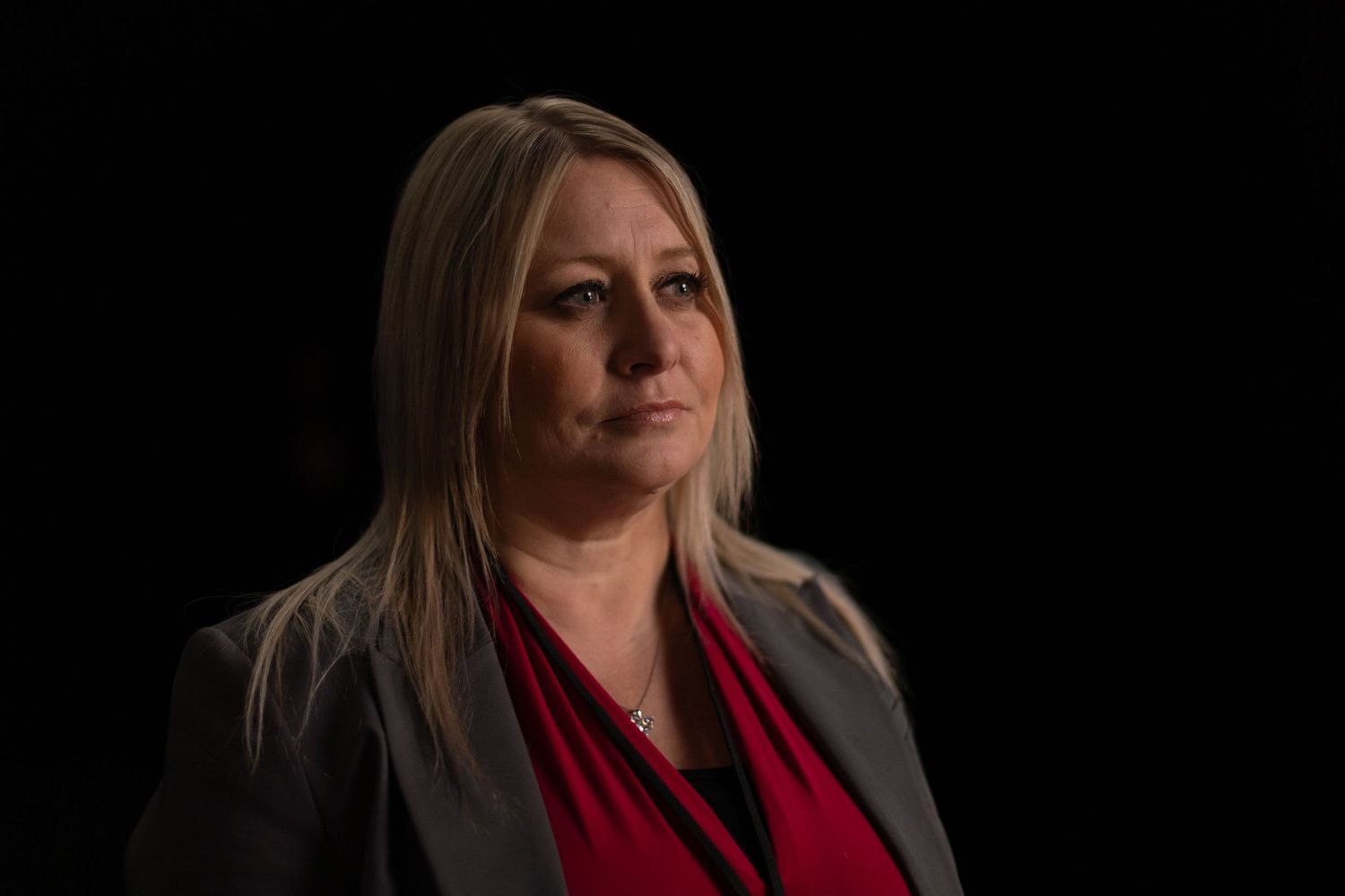 Professional headshot portrait with person wearing a black blazer and red top against dark background.