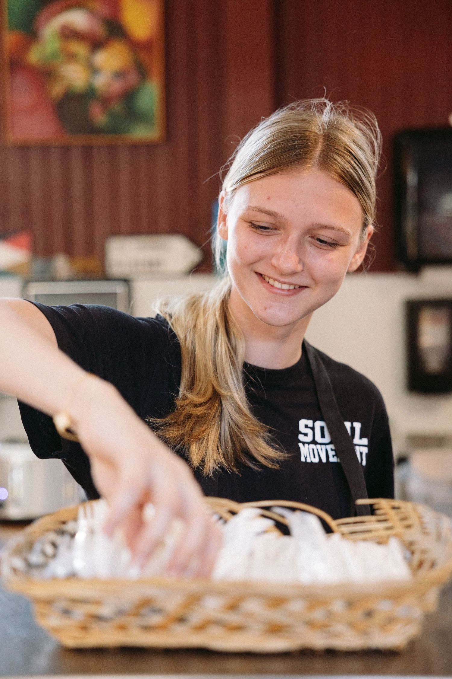 Server wearing black uniform reaches into wicker basket at restaurant counter while smiling warmly.