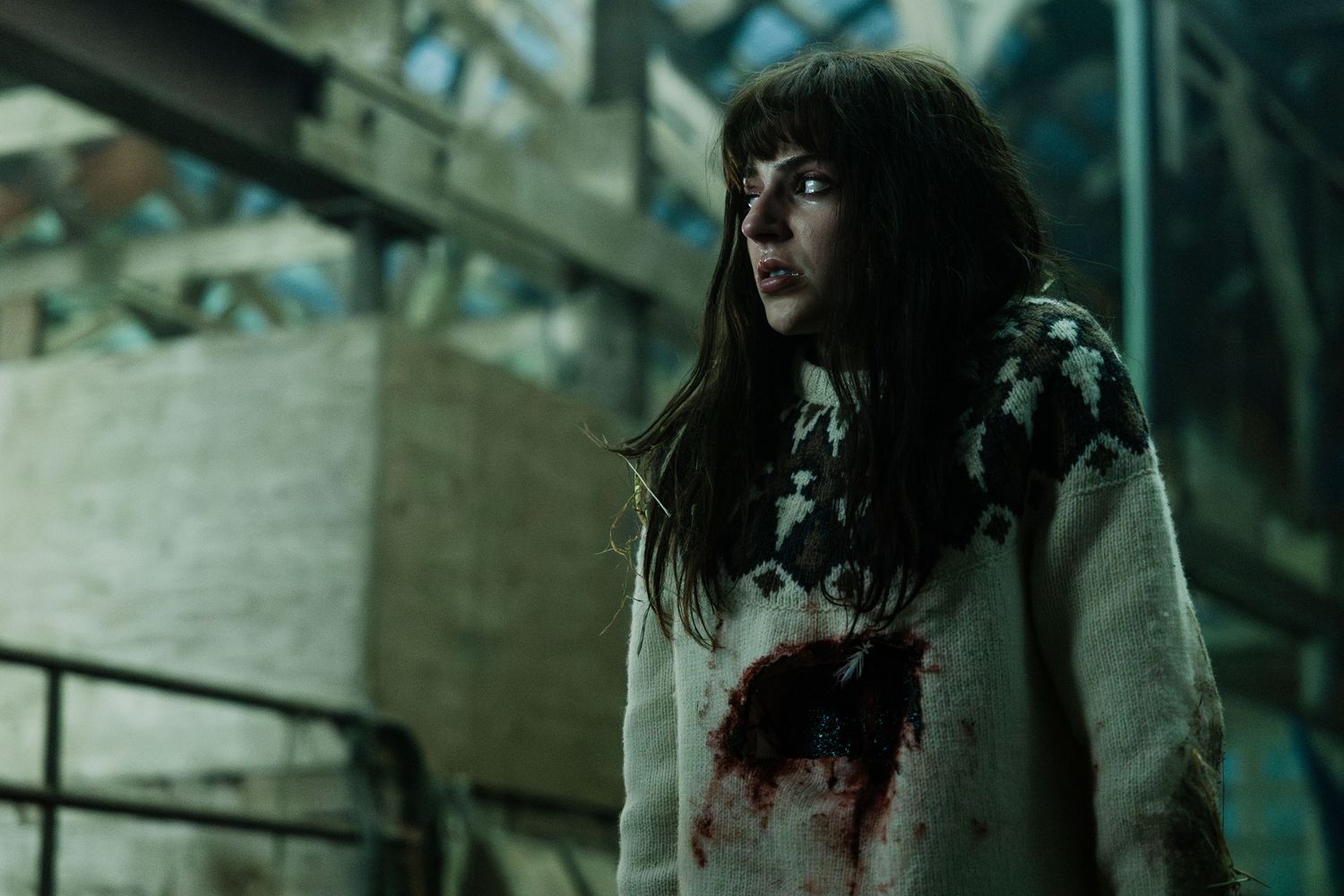 Someone in a blood-stained white sweater stands in an eerie industrial setting with dark lighting.