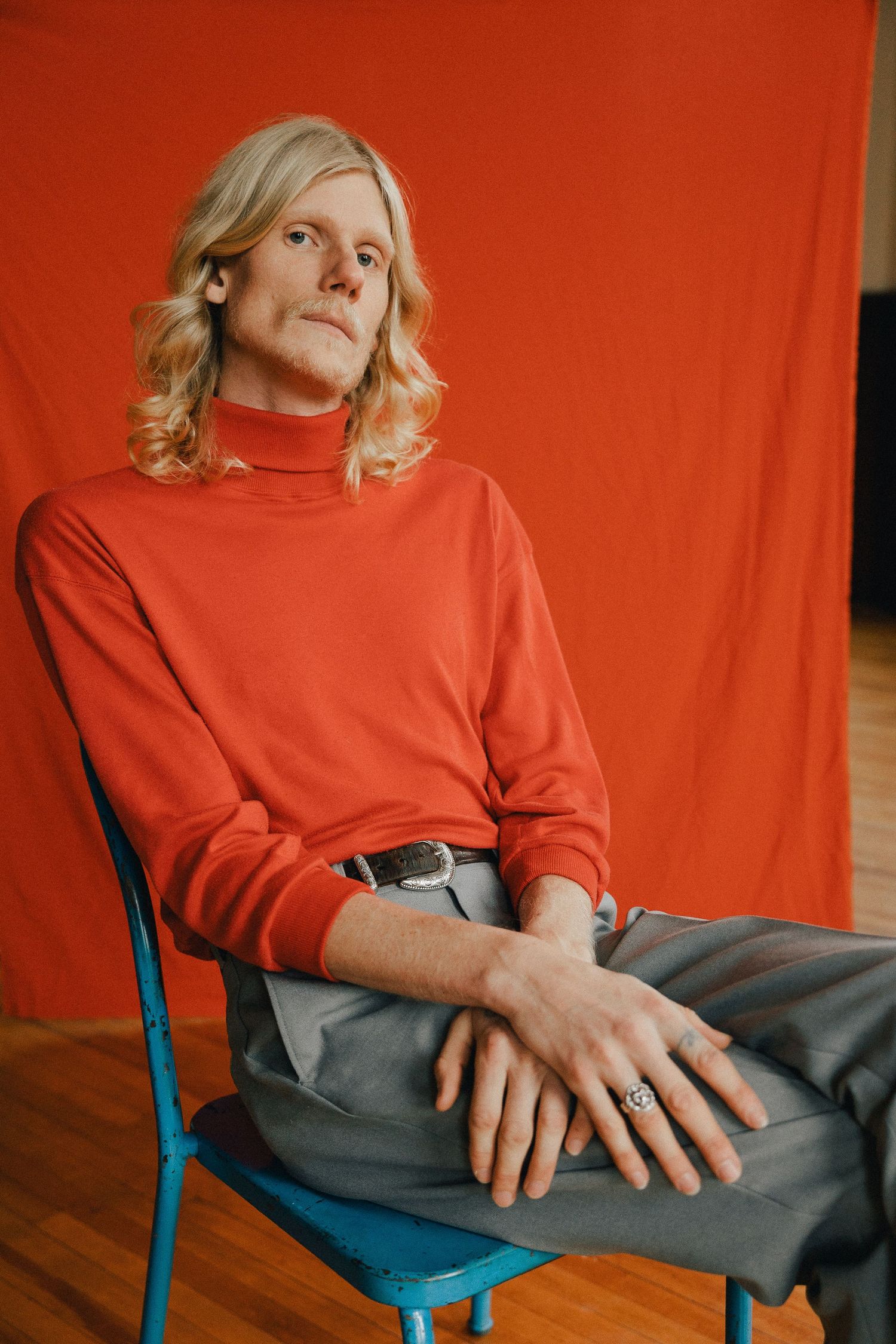 A person in a red turtleneck sweater sits on a turquoise chair against an orange backdrop with blonde wavy hair.