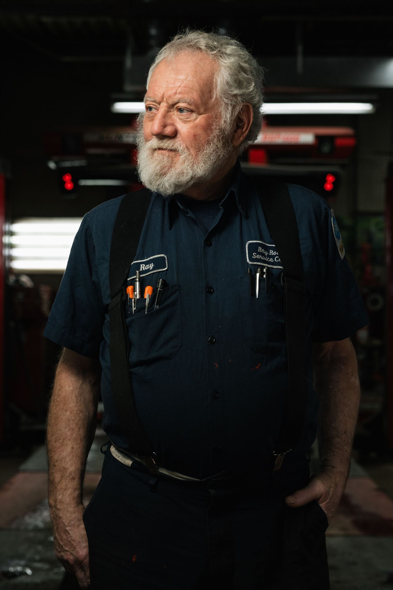Senior mechanic in navy blue work uniform with pens in pocket stands confidently in dimly lit auto repair shop.