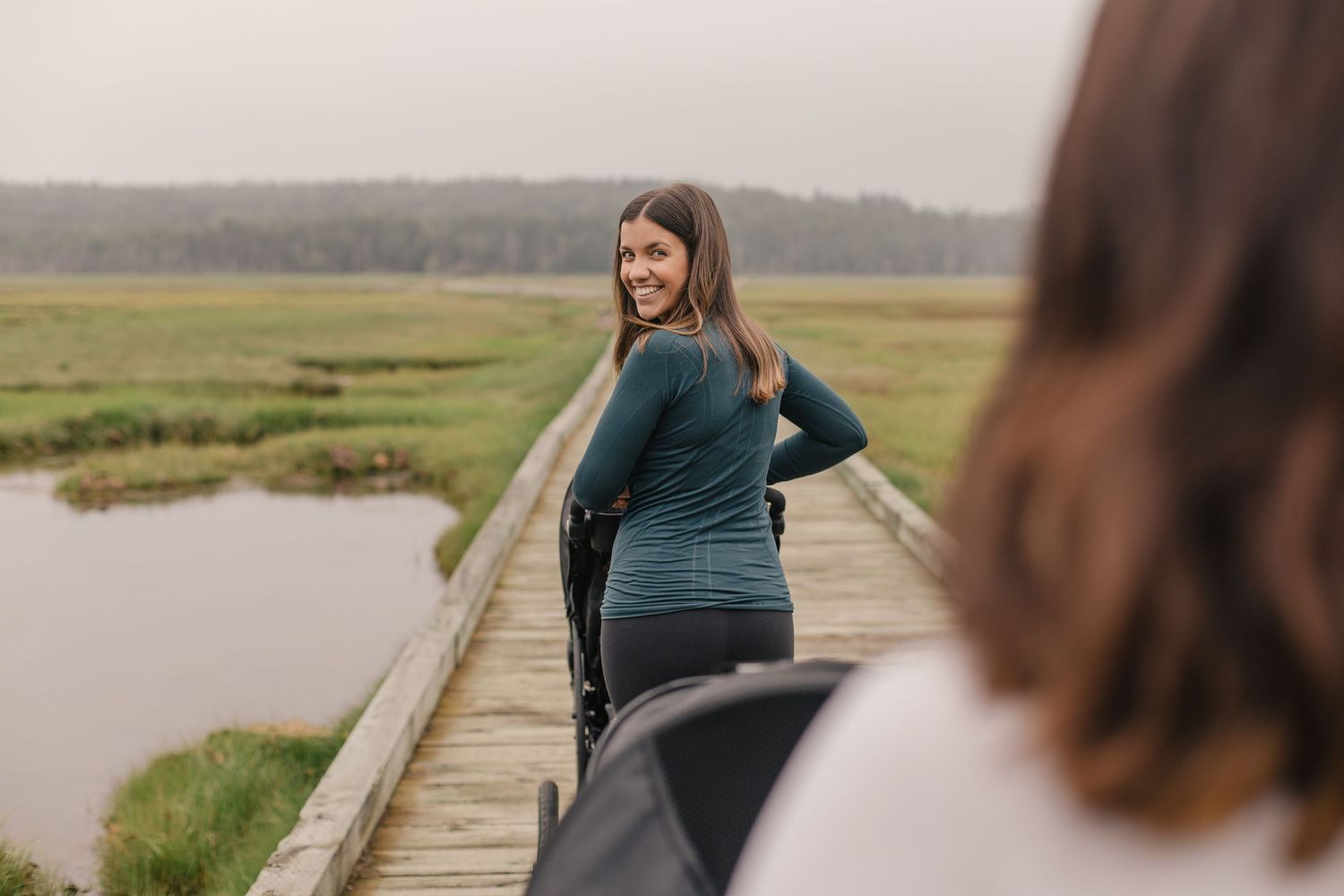 Two people walk on a wooden boardwalk through a marshy wetland landscape surrounded by grass and water.