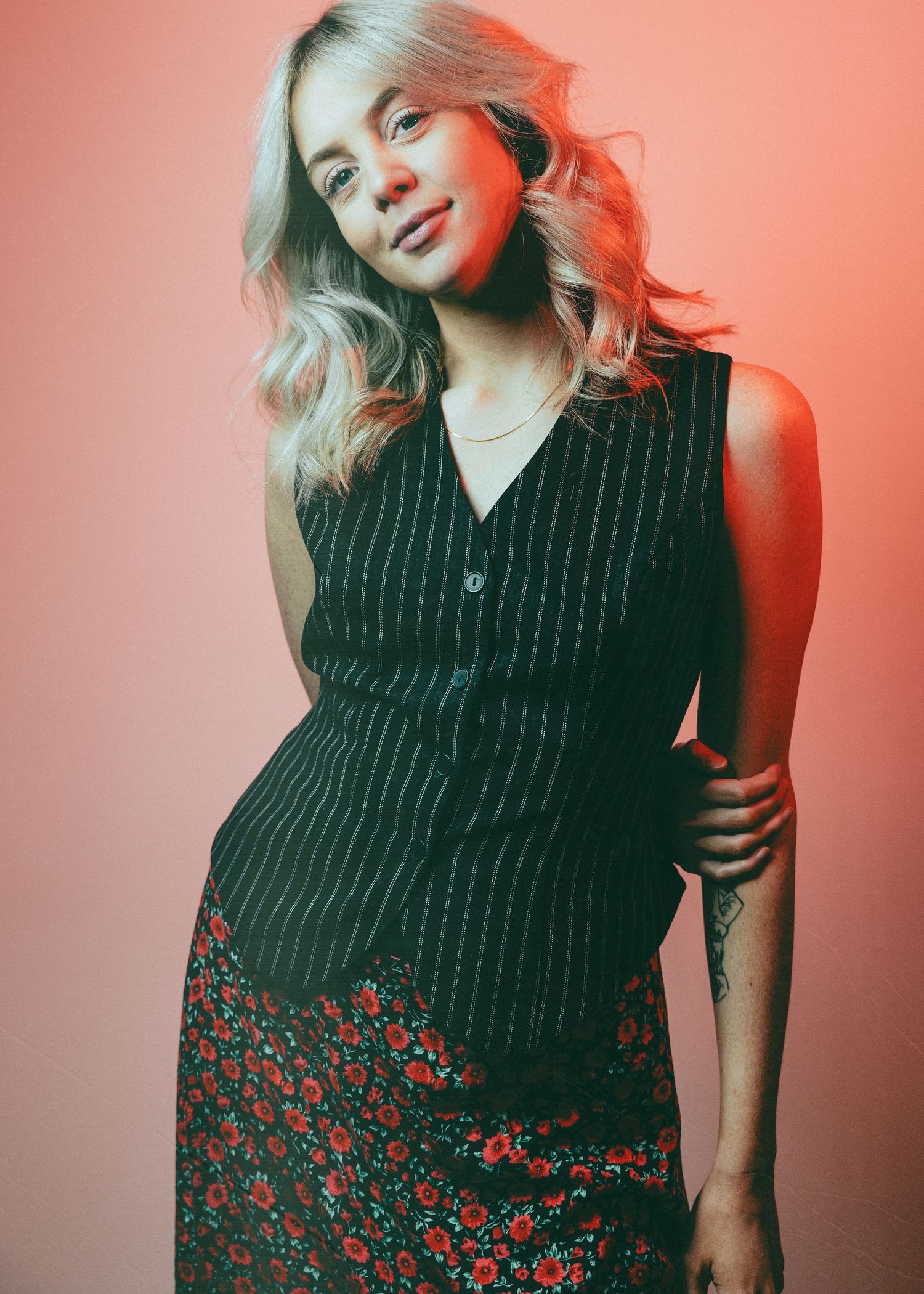 Stylish model in pinstripe vest and floral skirt poses against coral backdrop with soft lighting and wavy blonde hair.