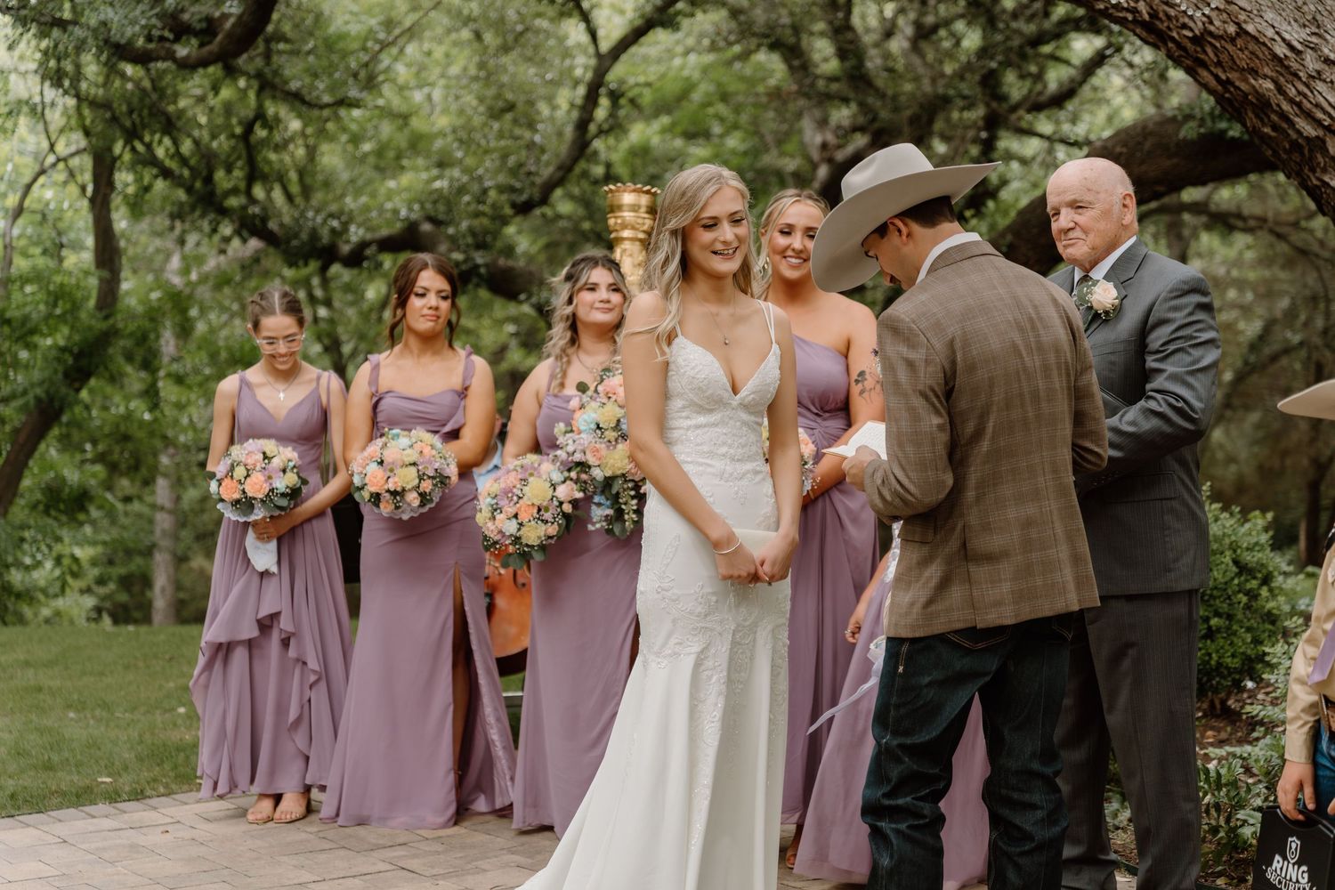 Bride in white gown with bridesmaids in lavender dresses holding floral bouquets at an outdoor wedding ceremony.