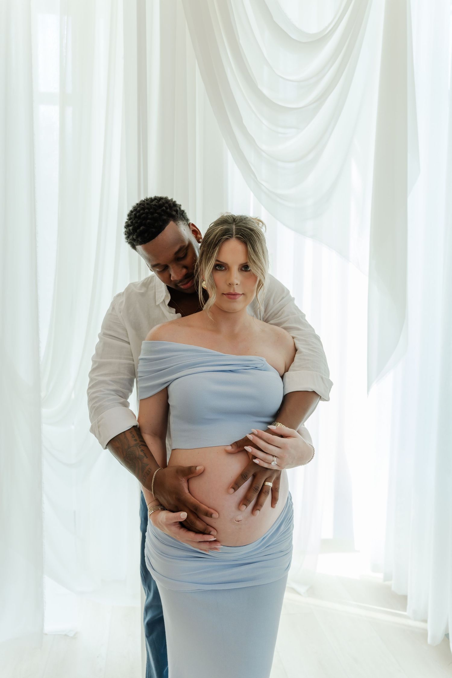A couple in light blue maternity attire embraces tenderly during a pregnancy photoshoot with white curtains in background.