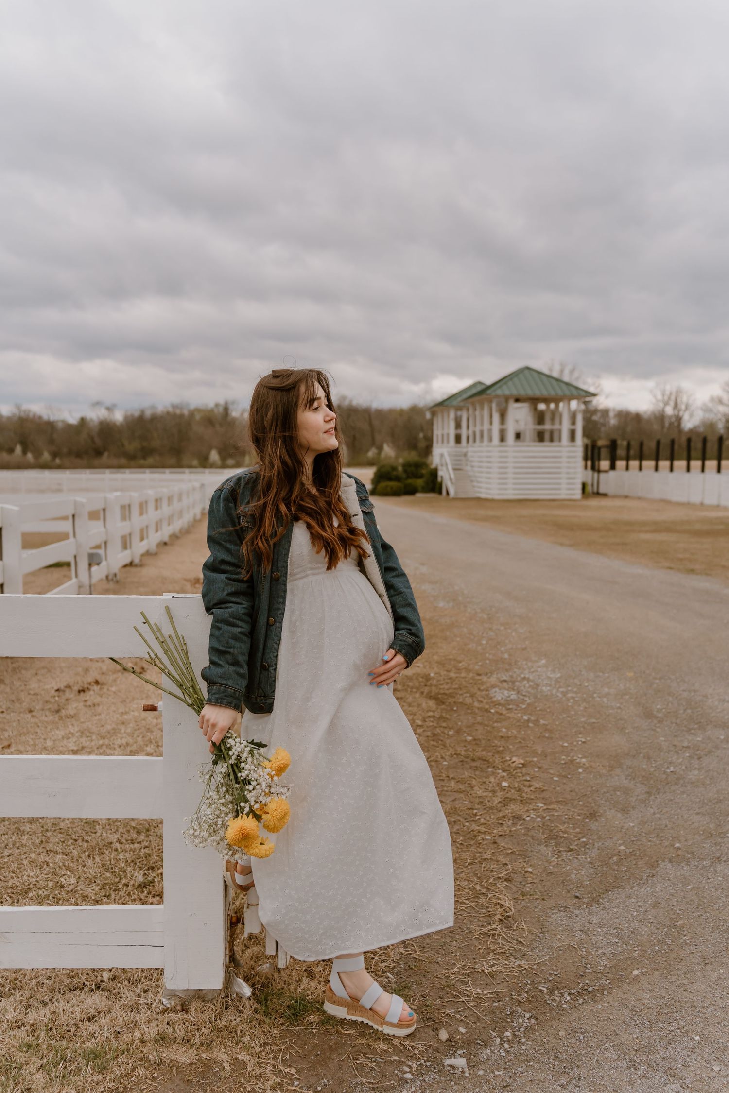 Figure in white dress and leather jacket holds yellow flowers while walking along a white fence.