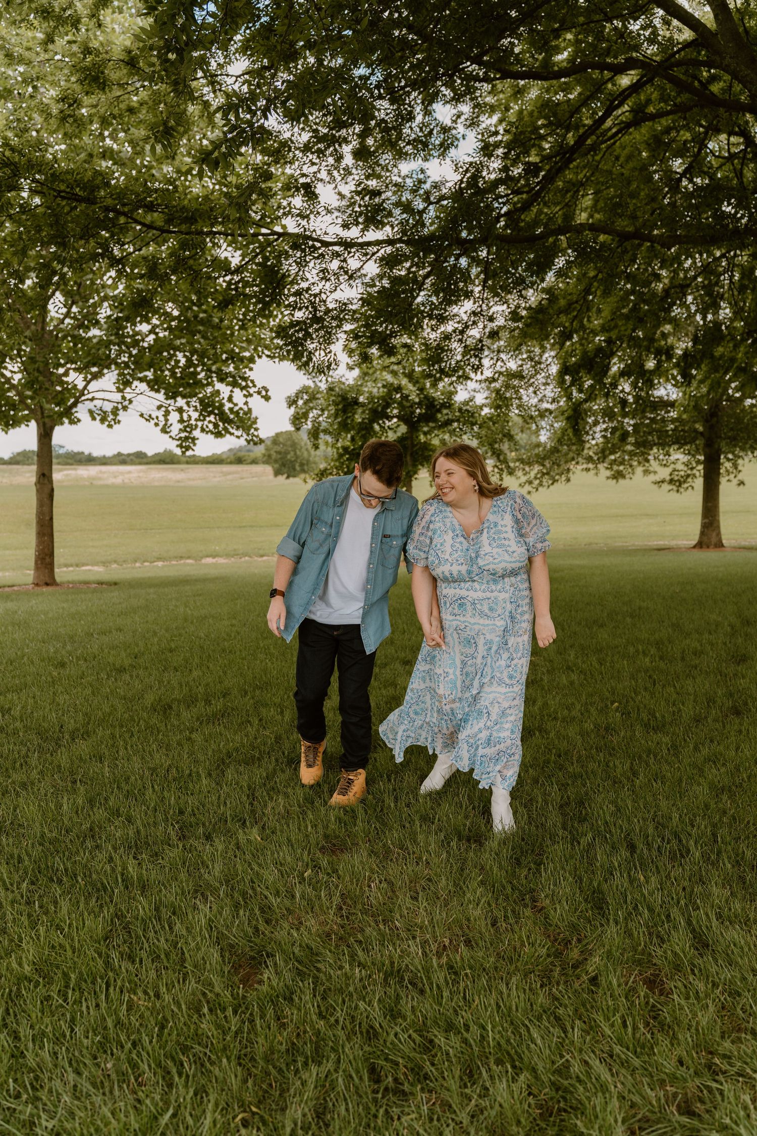 A couple walks together on a grassy lawn under large shade trees on a sunny afternoon.