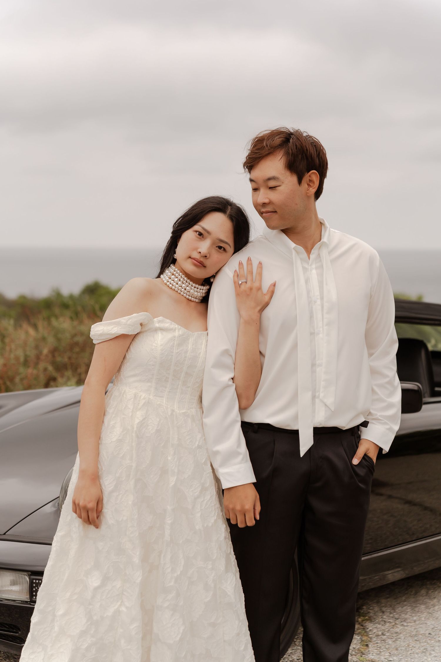 Asian couple in wedding attire pose by black car with ocean backdrop, bride in white gown with pearl choker.