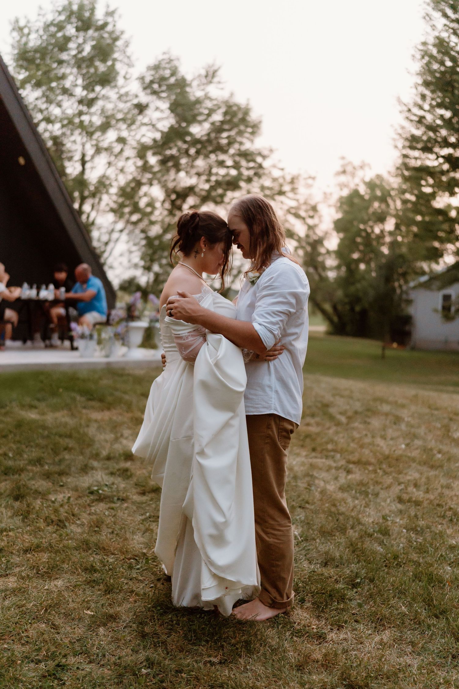 Couple sharing an intimate first dance on grass at outdoor wedding reception at dusk, bride in white gown.
