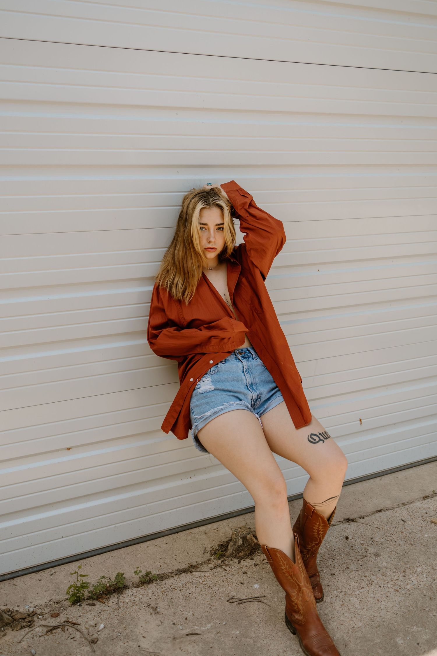 Casual fashion look with rust-colored jacket, denim shorts and brown cowboy boots against white garage door.