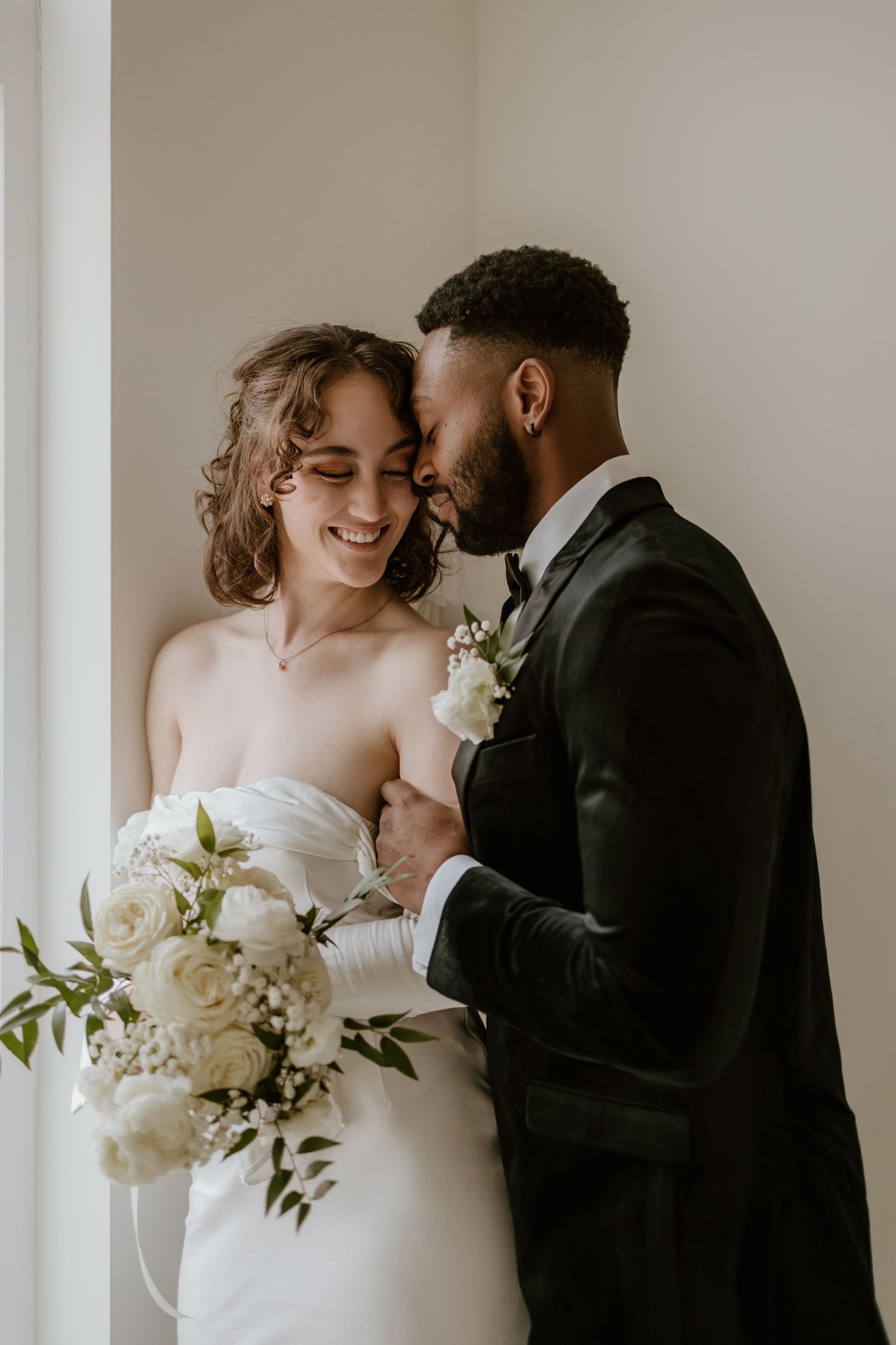 Romantic wedding couple embraces near window with white floral bouquet in elegant indoor setting.