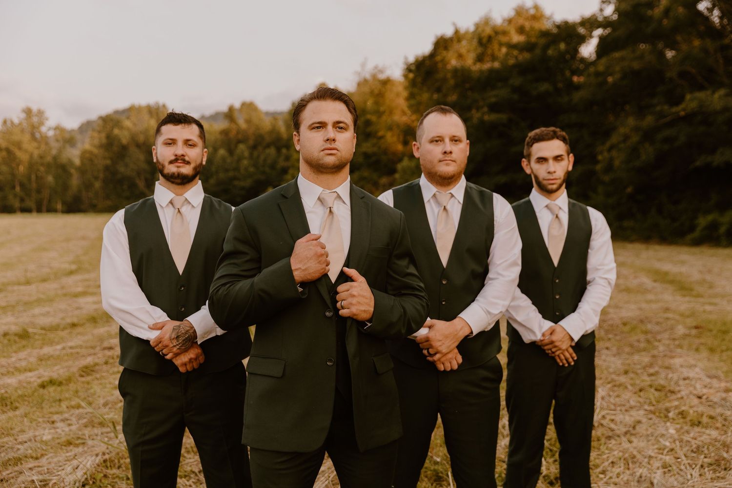 Groomsmen in dark suits and pink ties pose together outdoors at sunset.