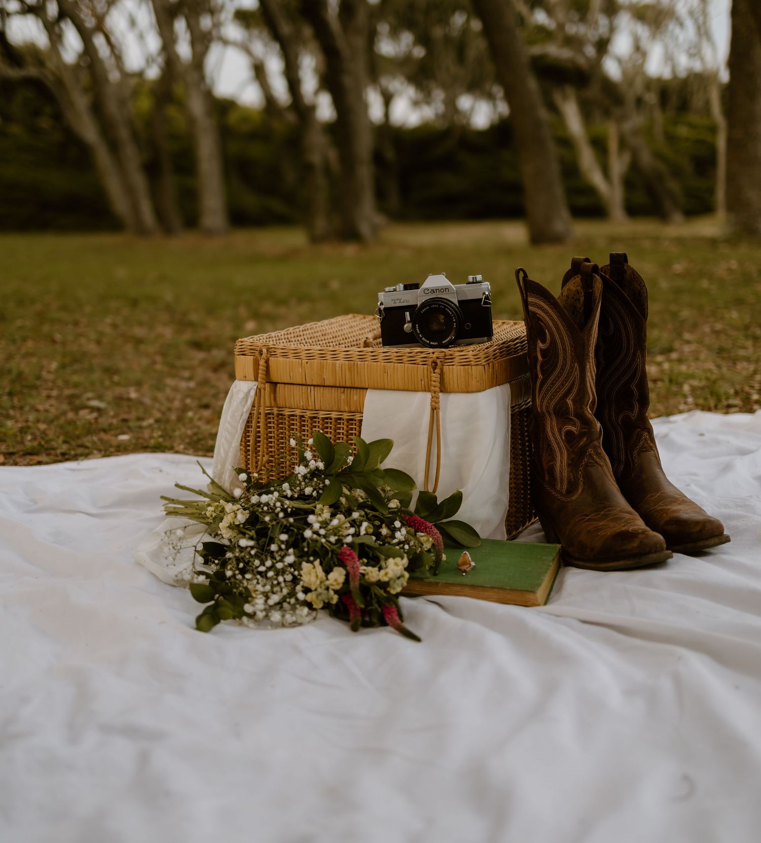 Vintage camera, cowboy boots, and wildflowers arranged on white blanket for styled photography.