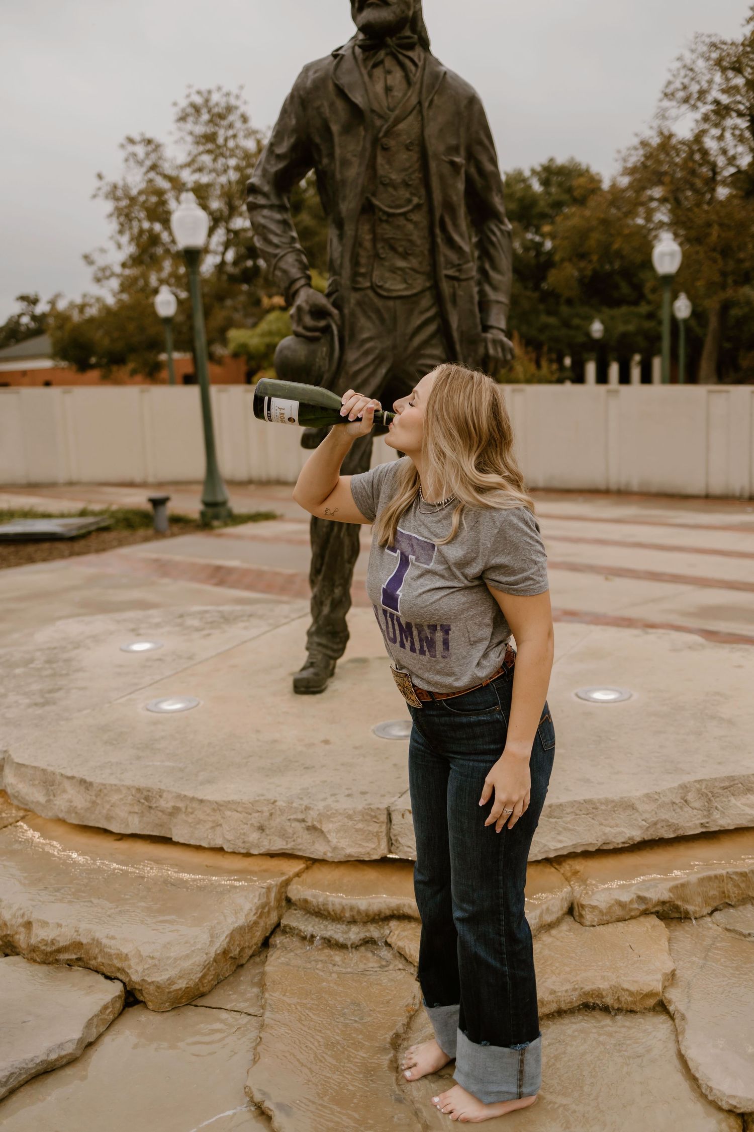 A playful photo shoot at a statue fountain shows someone drinking from a wine bottle in casual attire.
