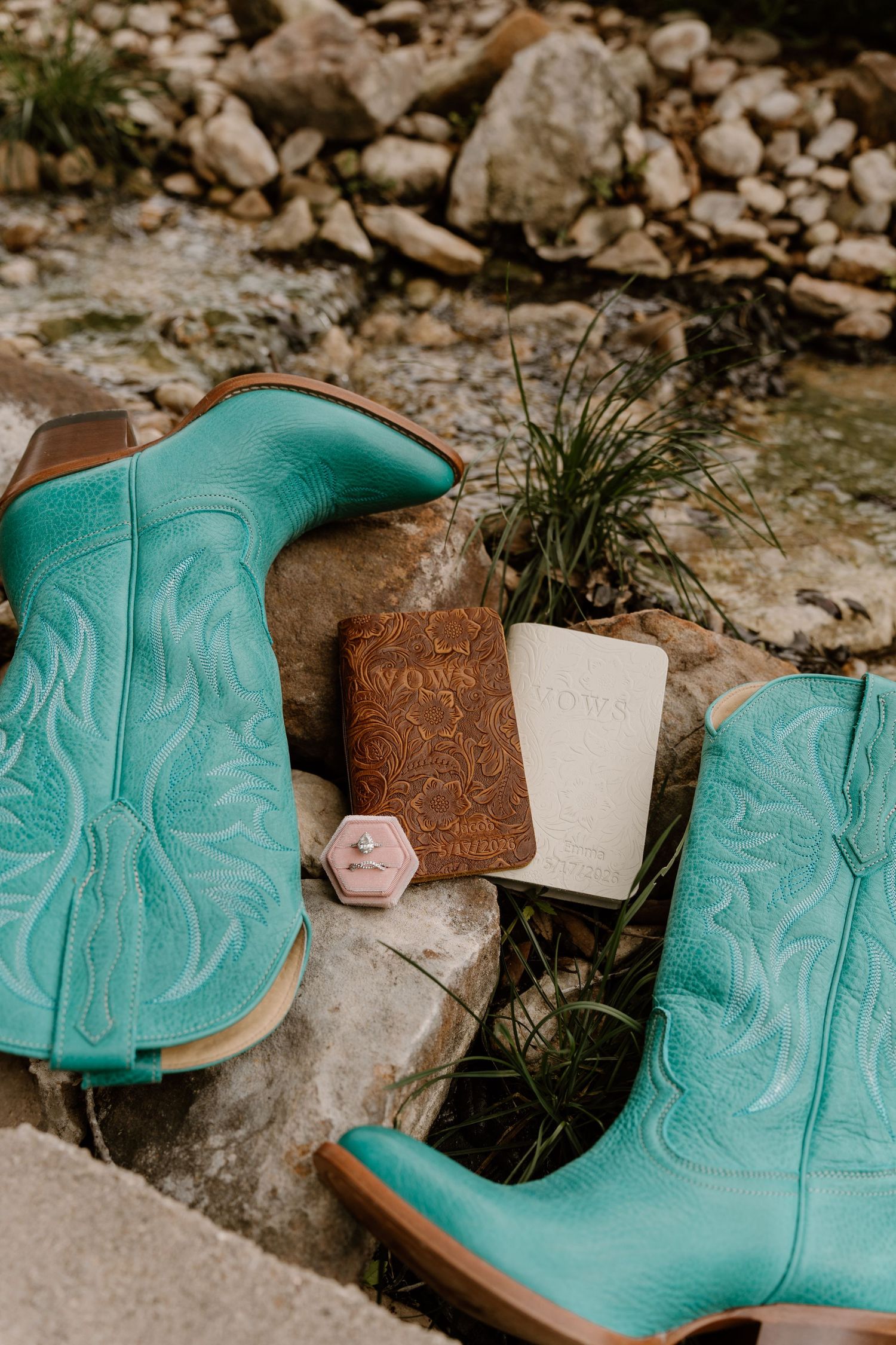 Turquoise cowboy boots with vow books and a pink ring box on rocky creek stones for a rustic western wedding.