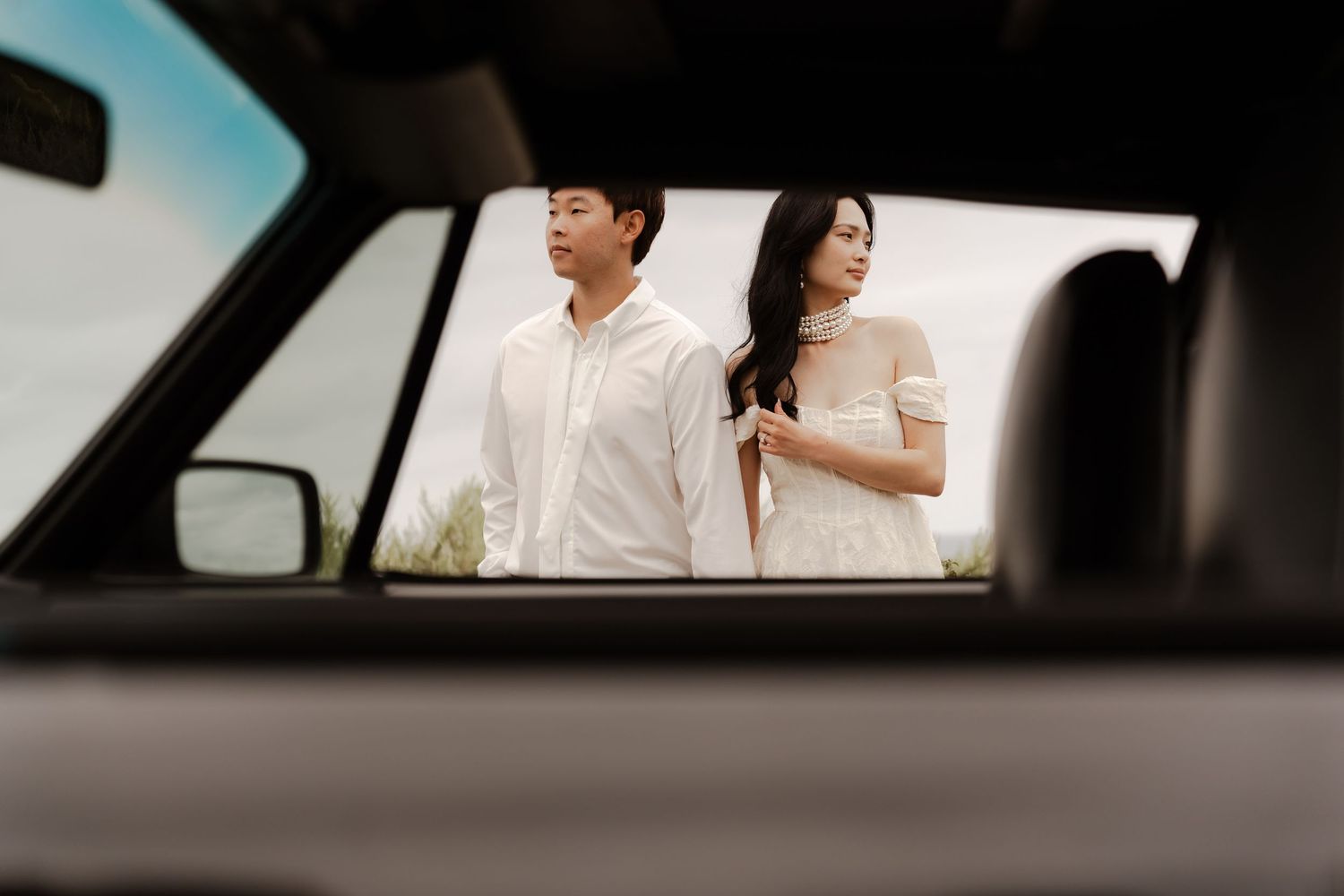 A couple in white attire stands outdoors, viewed through a car window in an elegant engagement photo.