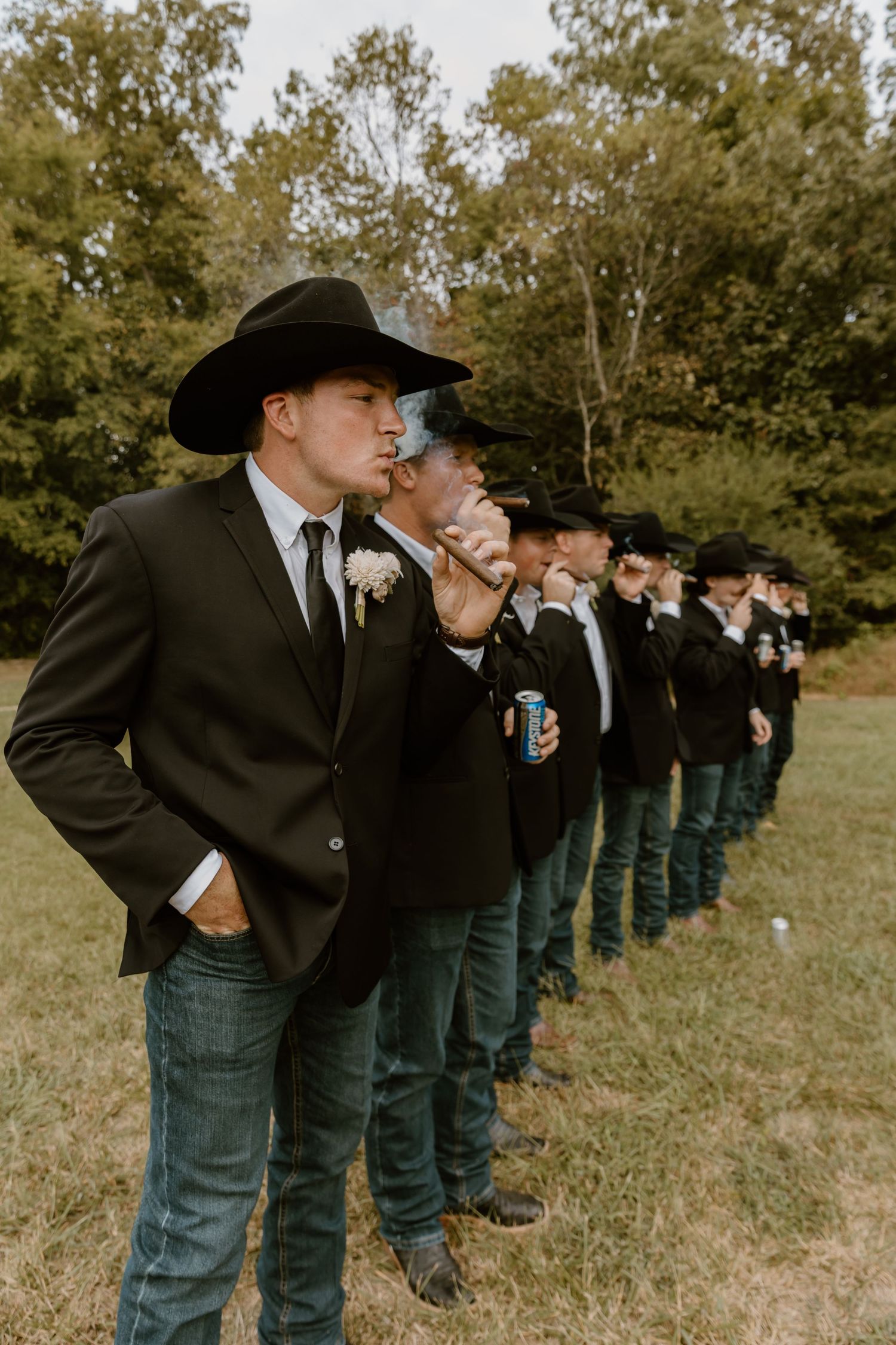 Groomsmen in black suits and cowboy hats lined up outdoors in autumn.