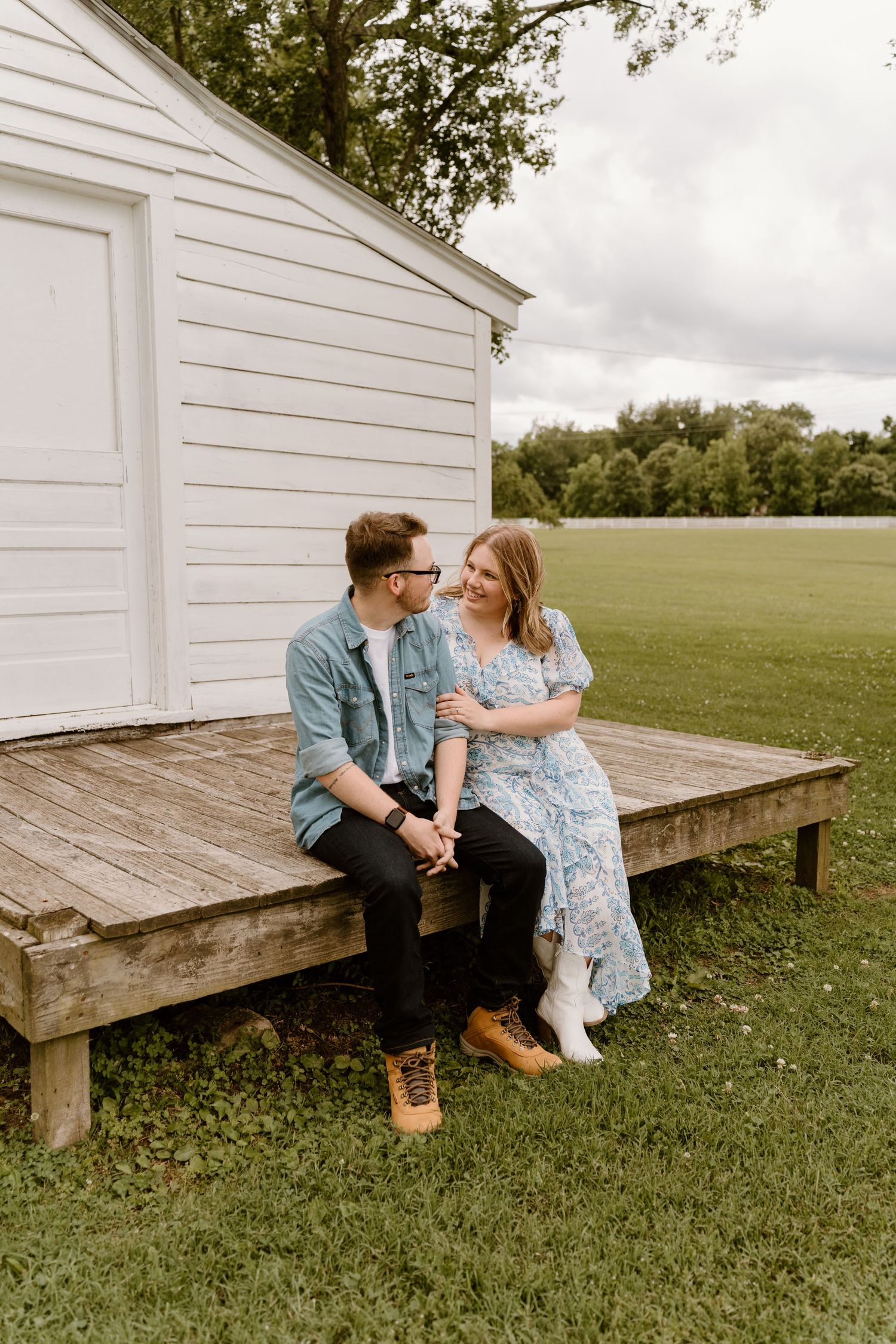 A couple sits on a wooden deck outside a rustic white farmhouse.