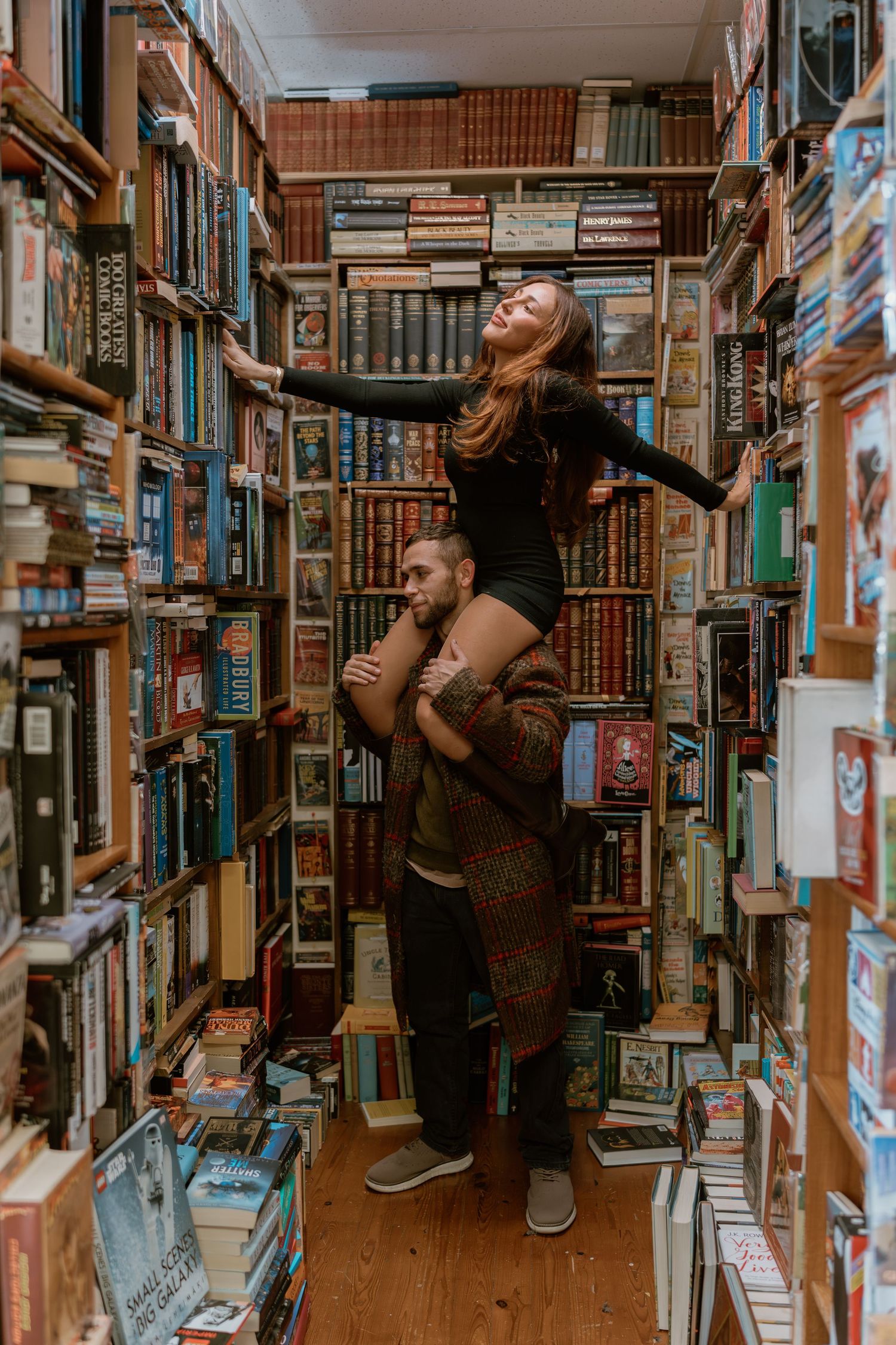 Person in black outfit browses books while doing creative dance poses in a cozy bookstore aisle.