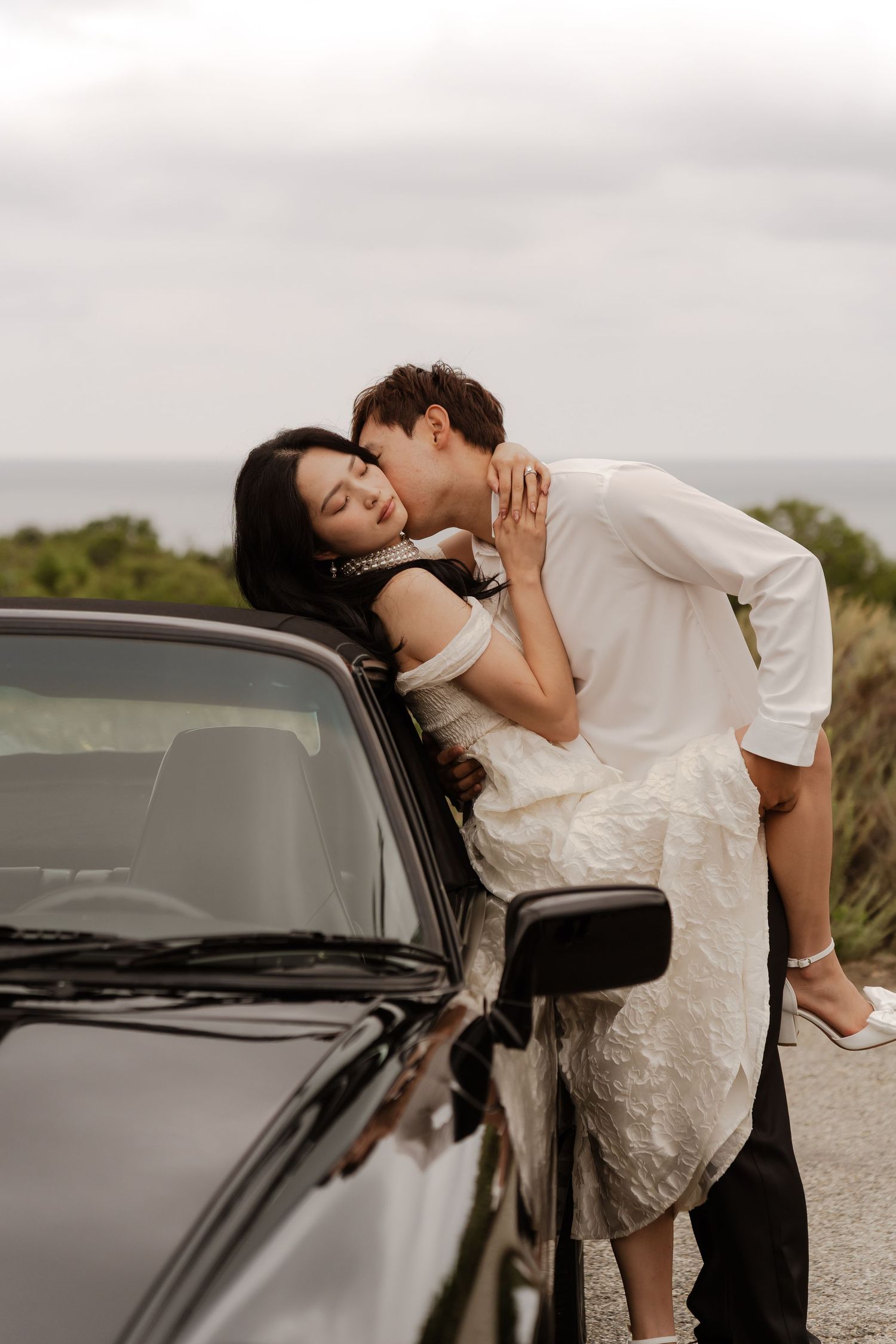 A couple in wedding attire shares a romantic kiss beside a black car with an ocean backdrop.